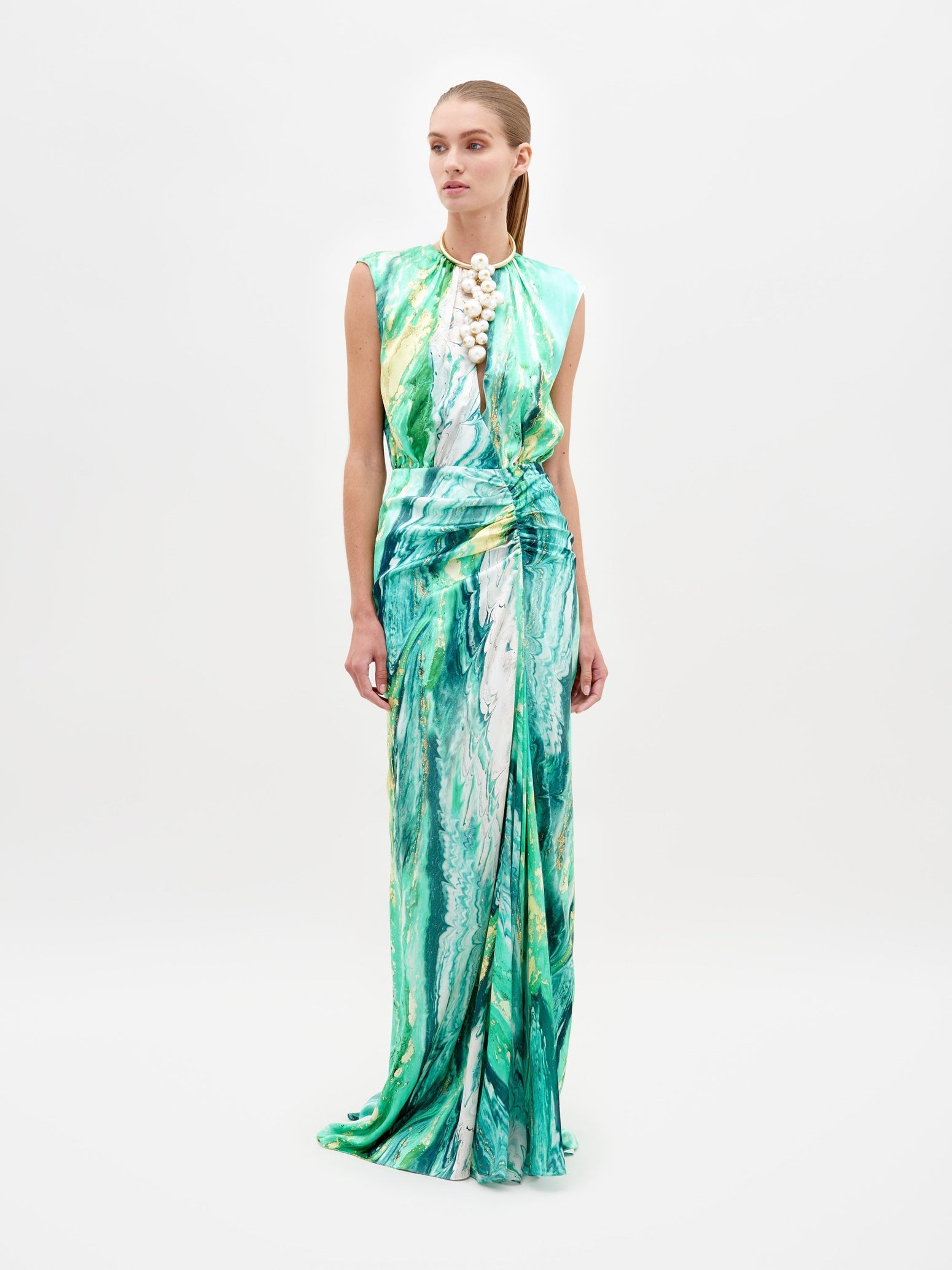 A person stands wearing the Samaira Dress Green Marble, a sleeveless, floor-length design featuring a striking green and white marble pattern. The outfit is completed with a high necklace adorned with large white beads, set against a plain white background. Pre-order now to ensure you receive yours by the February 15th, 2025 ship date.