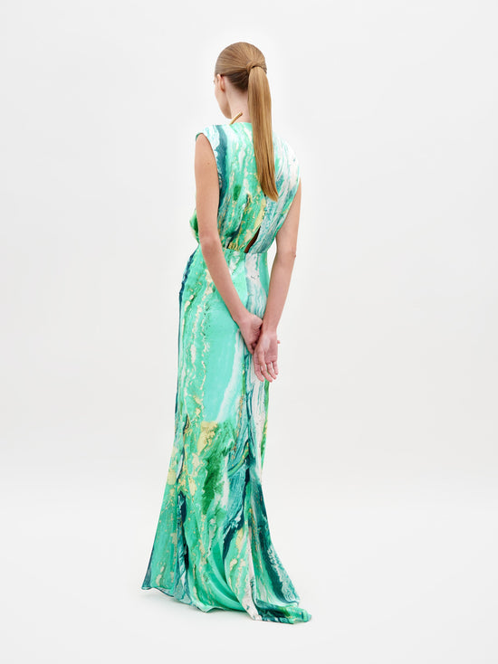 A person stands wearing the Samaira Dress Green Marble, a sleeveless, floor-length design featuring a striking green and white marble pattern. The outfit is completed with a high necklace adorned with large white beads, set against a plain white background. Pre-order now to ensure you receive yours by the February 15th, 2025 ship date.