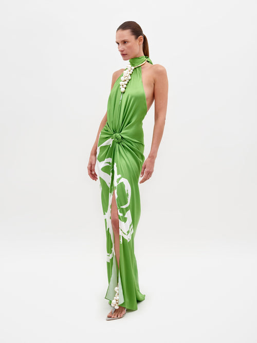 A woman wearing the Olatz Dress Green Bloom, featuring a green halter neck design with white floral accents and a front knot detail, stands against a plain background. Available for pre-order now; secure your stylish look today. Ships by 2025.