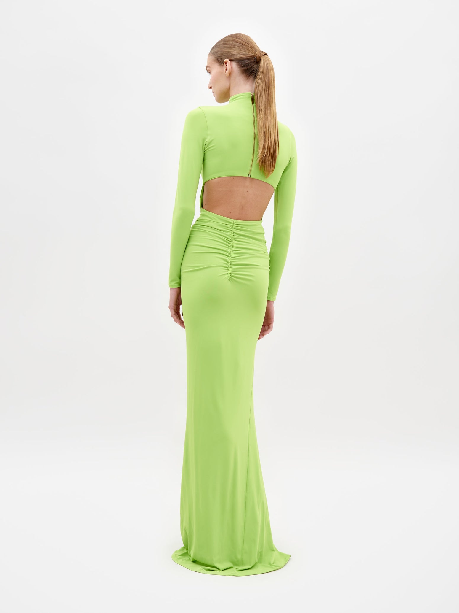 A person is wearing the Lena Dress Lime, a long, lime green dress with long sleeves and a high neckline. The dress, which is available for shipping by February 15th, features cut-out details at the sides and a gathered, draped front. White background.