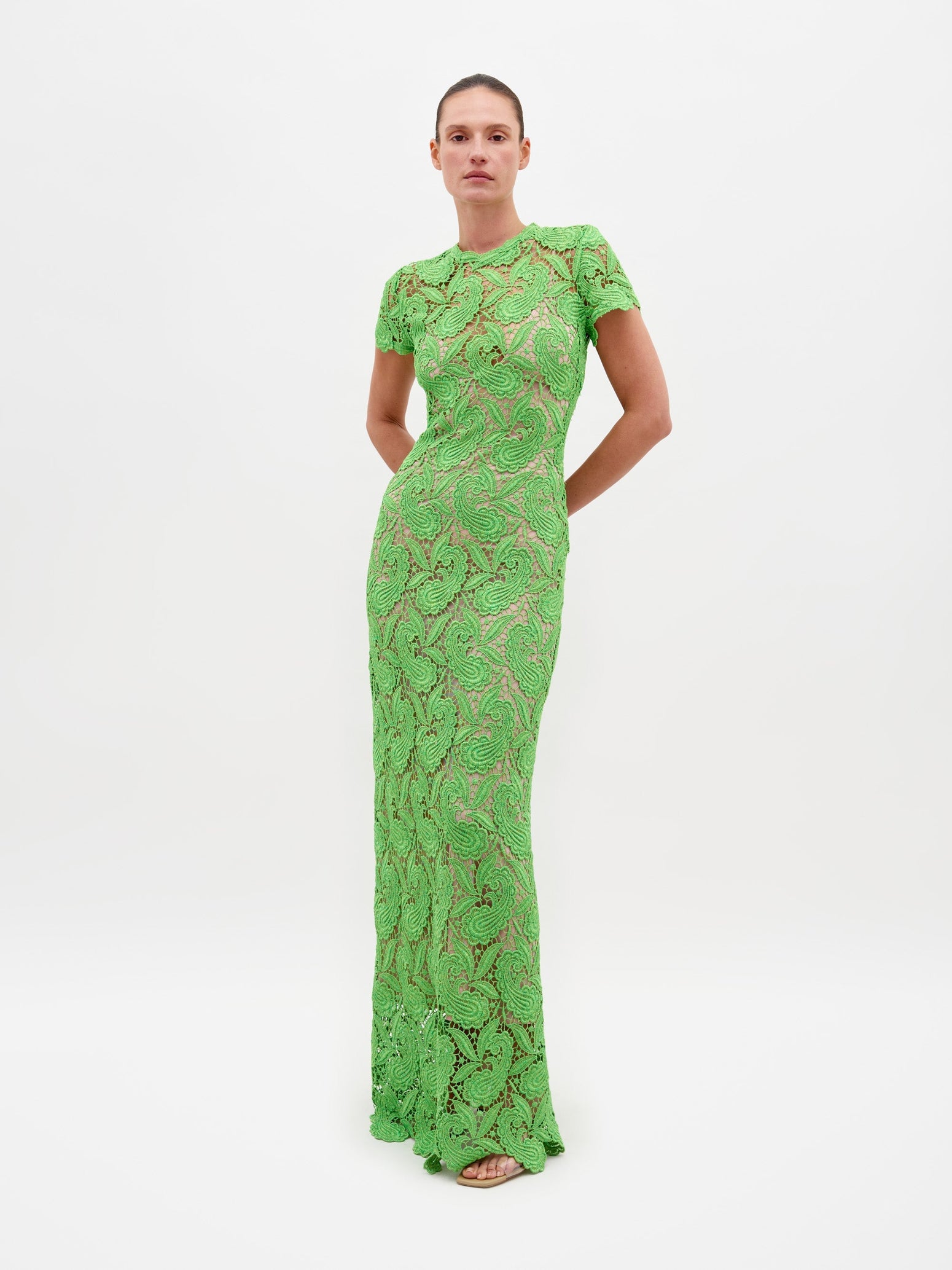 A person stands against a plain white background, wearing the Roxana Dress Green Guipure—a long, fitted lace dress with short sleeves. Hands are clasped behind the back. This elegant dress is available for pre-order now, with a scheduled ship date of February 15th, 2025.