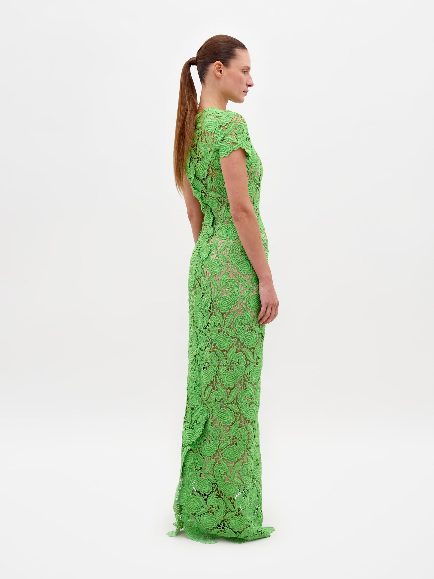 A person stands against a plain white background, wearing the Roxana Dress Green Guipure—a long, fitted lace dress with short sleeves. Hands are clasped behind the back. This elegant dress is available for pre-order now, with a scheduled ship date of February 15th, 2025.