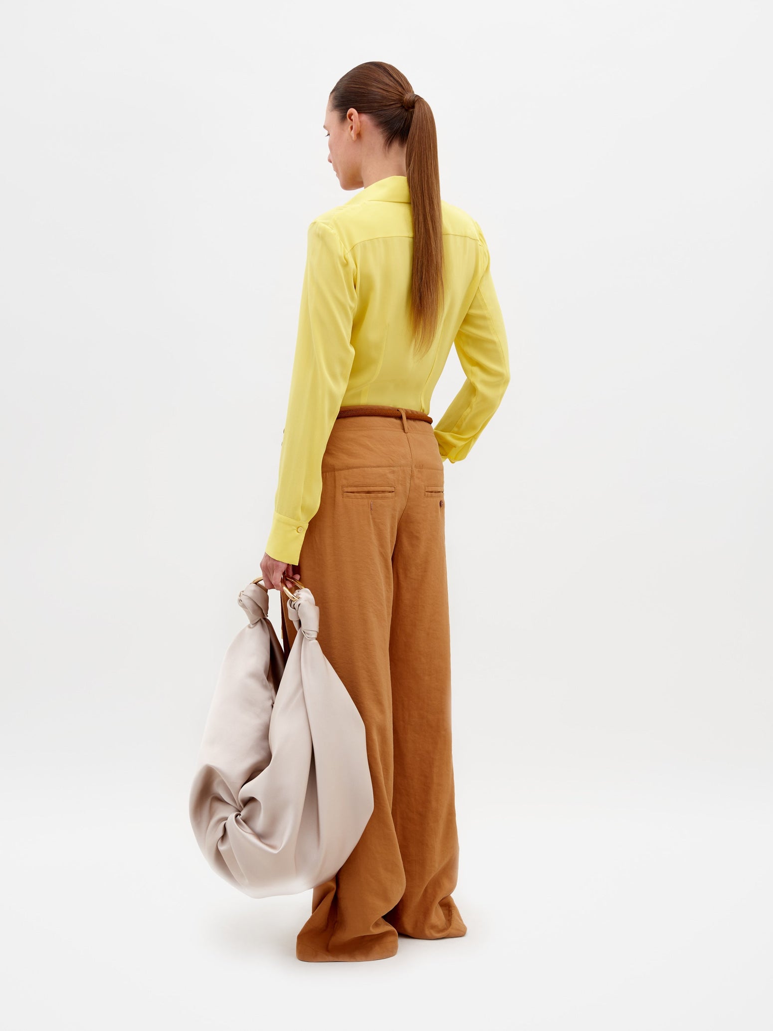 A person stands against a plain background wearing a yellow blouse, the Opal Pant Camel, and sandals, holding a large beige bag. Don't miss the chance to pre-order this outfit by February 15th, 2025.