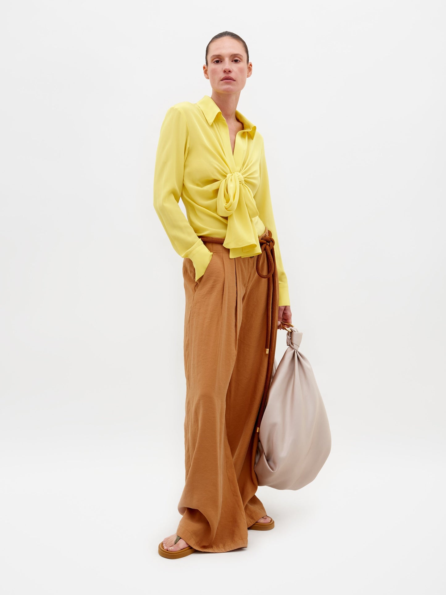 A person stands against a plain background wearing a yellow blouse, the Opal Pant Camel, and sandals, holding a large beige bag. Don't miss the chance to pre-order this outfit by February 15th, 2025.