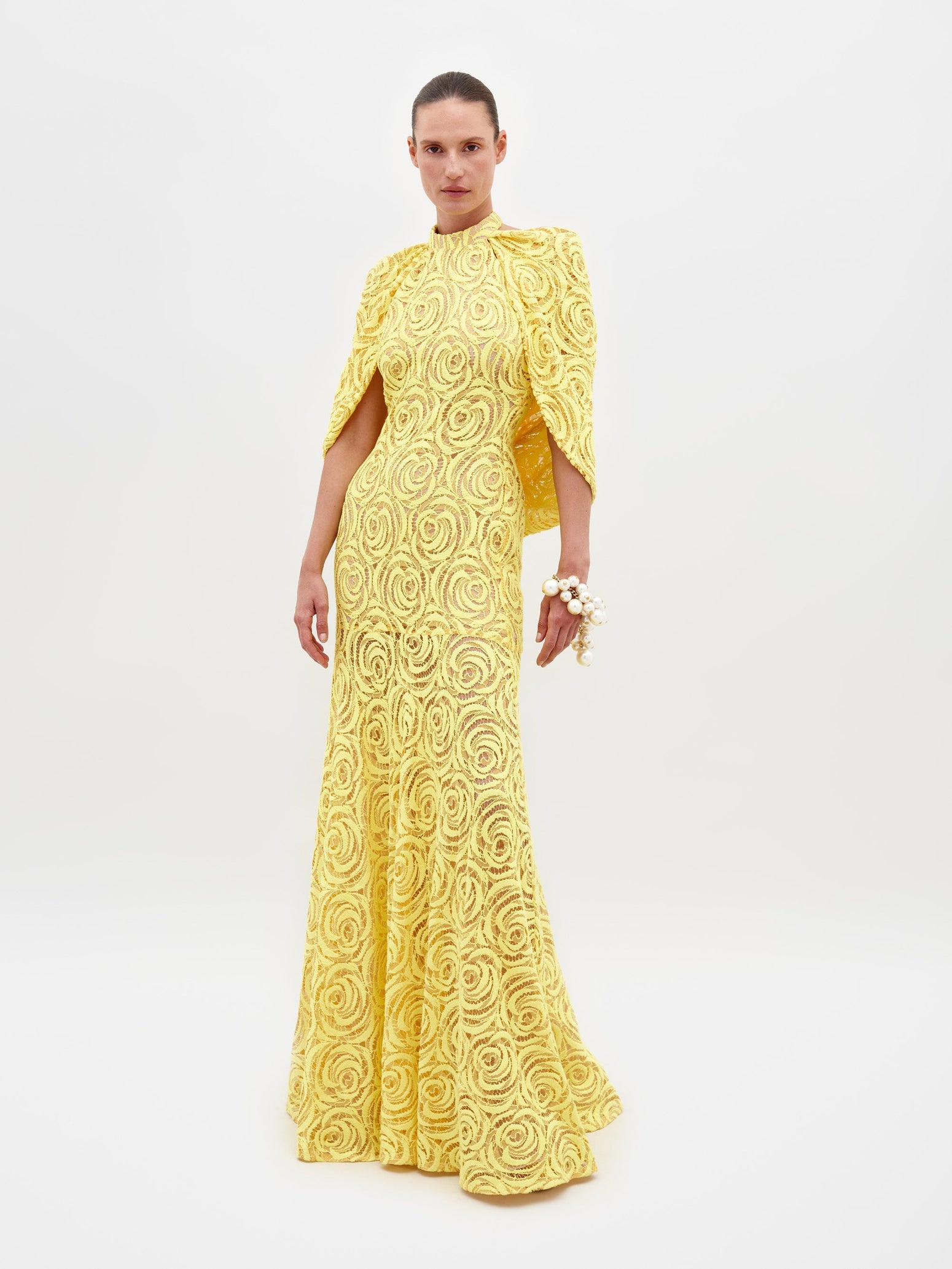 Person wearing a Salvina Dress Tuscany Yellow, adorned with a high neckline and cape-like sleeves, holding a pearl bracelet on their wrist, eagerly awaiting the pre-order ship date for 2025.