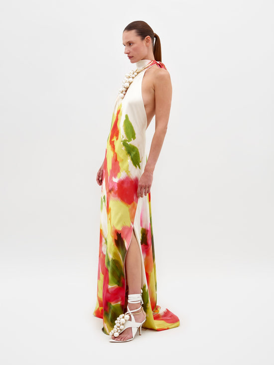 A person in the Sherry Dress Spring Blooms, featuring a multicolored print and a thigh-high slit, poses against a plain background. They are wearing white high-heeled sandals and a pearl necklace. Remember, this stunning outfit is available for pre-order and will ship by February 15th.