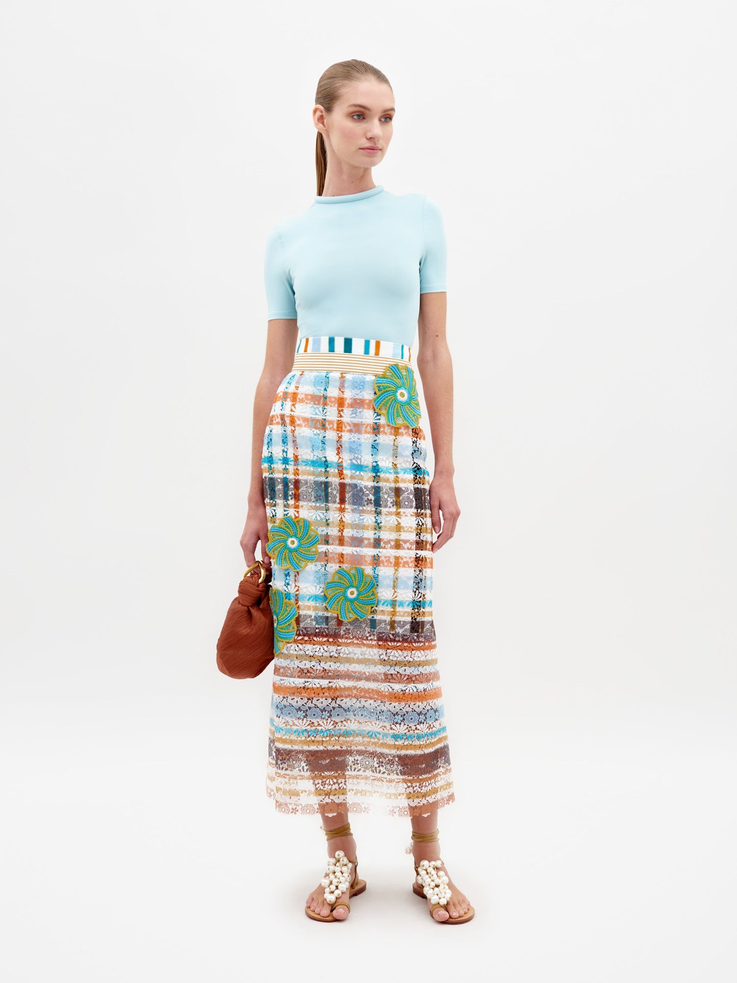 A woman stands against a plain background wearing a light blue top, the Shadia Skirt Multi Color Stripes, and white sandals. She holds a small brown bag, ready for her February 15th event in 2025.