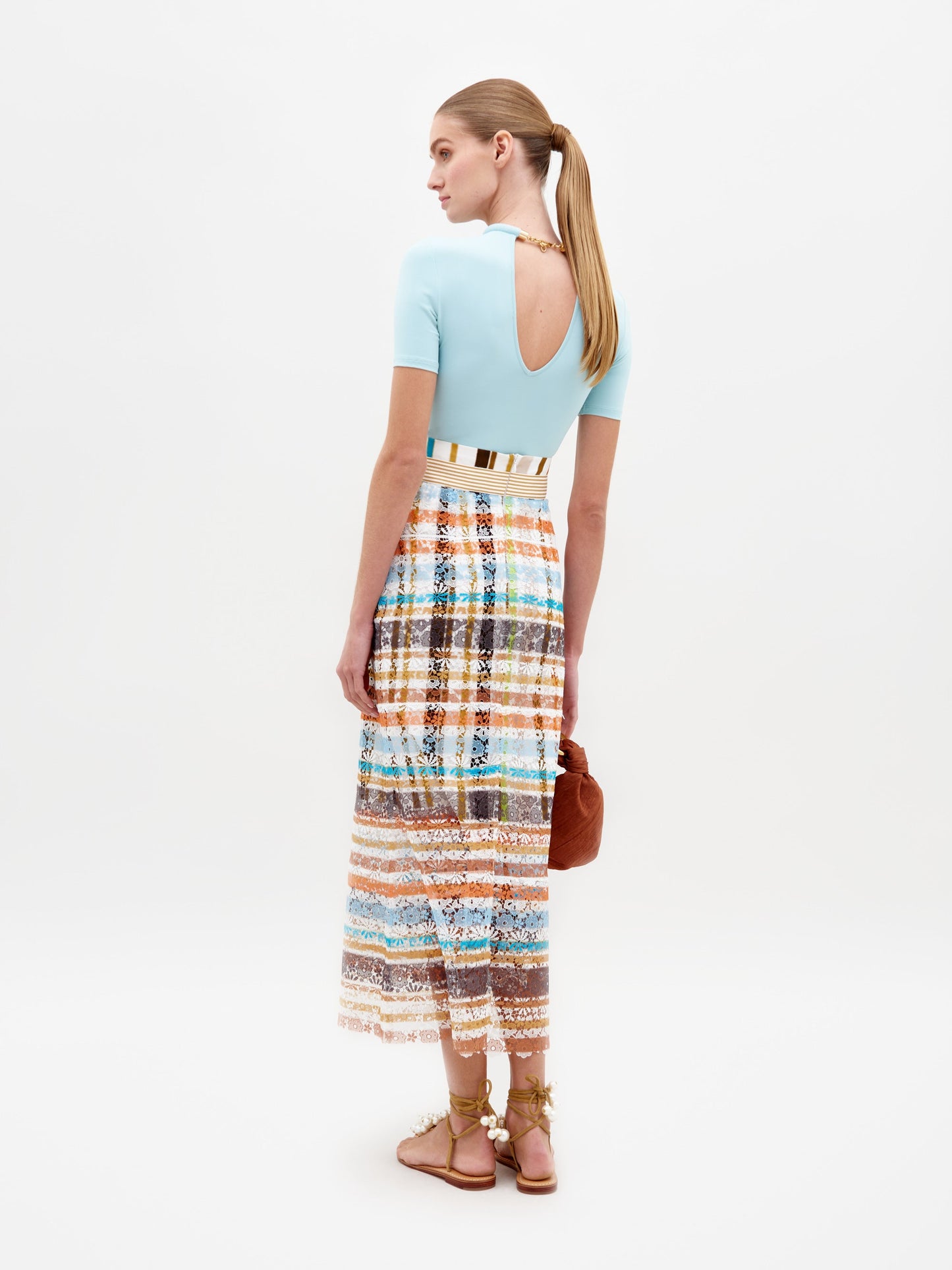 A woman stands against a plain background wearing a light blue top, the Shadia Skirt Multi Color Stripes, and white sandals. She holds a small brown bag, ready for her February 15th event in 2025.