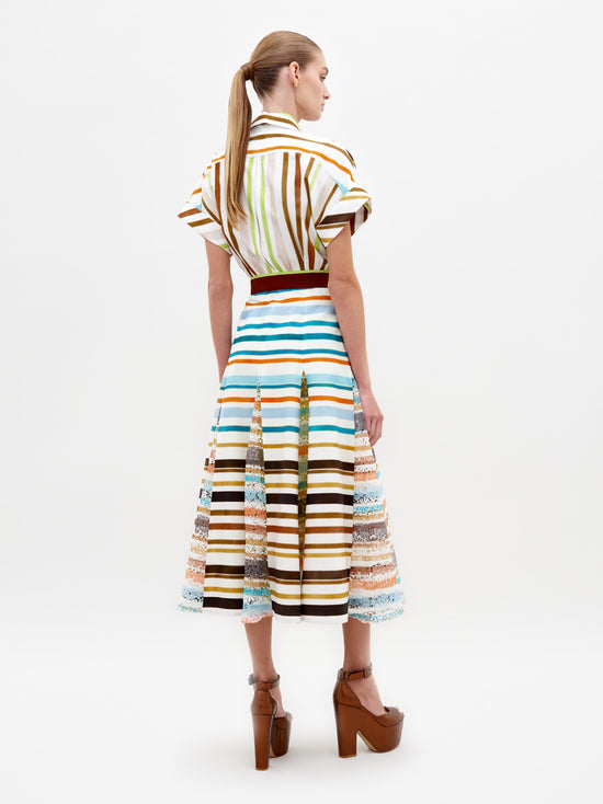 A model stands wearing the Odila Dress Multi Color Stripes paired with brown platform heels. The dress features vertical and horizontal stripes with a belted waist. This stylish ensemble, available for pre-order, will ship by early 2025. The background is plain white.