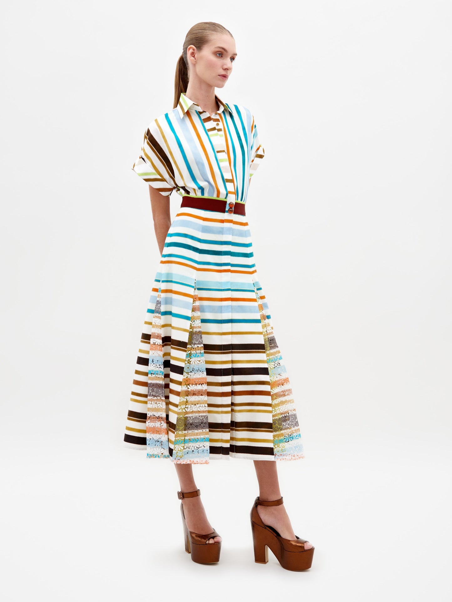 A model stands wearing the Odila Dress Multi Color Stripes paired with brown platform heels. The dress features vertical and horizontal stripes with a belted waist. This stylish ensemble, available for pre-order, will ship by early 2025. The background is plain white.