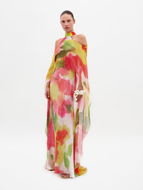 A person is standing in the Serenela Dress Spring Blooms, a colorful, flowing dress with a halter neck and floral patterns, holding a small white flower bouquet. The dress is ready to be pre-ordered for the February 15th ship-by date.