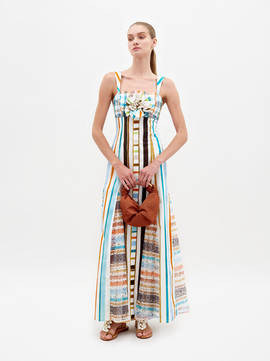 A woman stands wearing the Ofra Dress Multi Color Stripes, a sleeveless outfit featuring vibrant stripes and floral details, while holding an orange bag. The background is plain white. Please note this item is available for pre-order and will ship by February 15th, 2025.