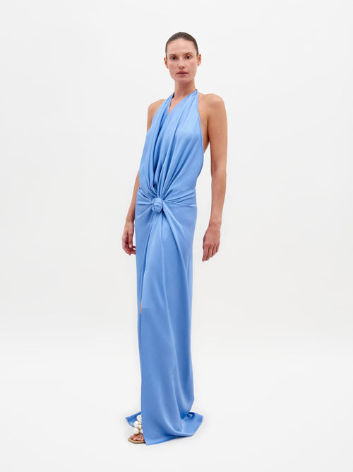 A person in the Torgiano Dress Periwinkle, a stunning blue halter-neck design with a central knot and an elongated hem, stands against a plain white background. This elegant piece is available for pre-order now and will ship by February 15th, 2025.