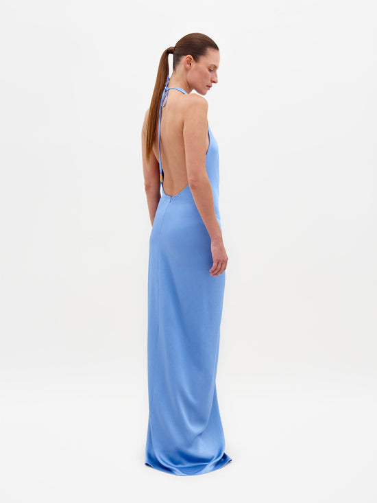 A person in the Torgiano Dress Periwinkle, a stunning blue halter-neck design with a central knot and an elongated hem, stands against a plain white background. This elegant piece is available for pre-order now and will ship by February 15th, 2025.