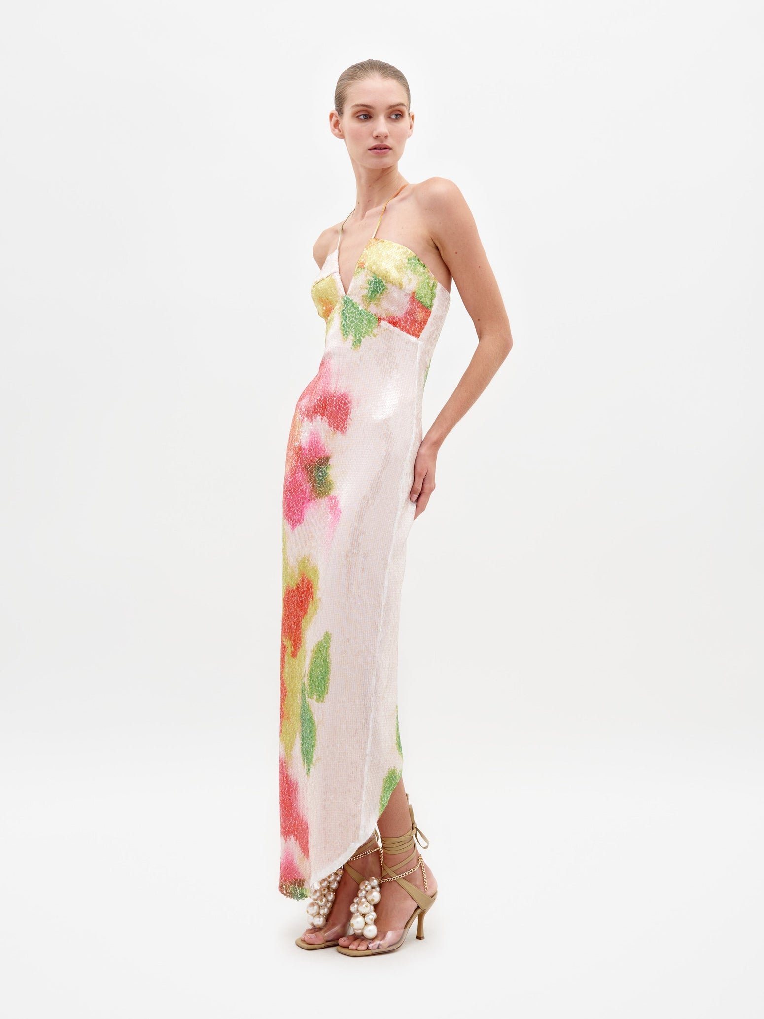 A woman is wearing the Lisseth Dress Spring Blooms, a strapless white dress adorned with colorful floral patterns, paired with high-heeled sandals featuring ankle straps and large pearl embellishments—an ideal choice for those who pre-order by February 15th to ensure timely shipping.