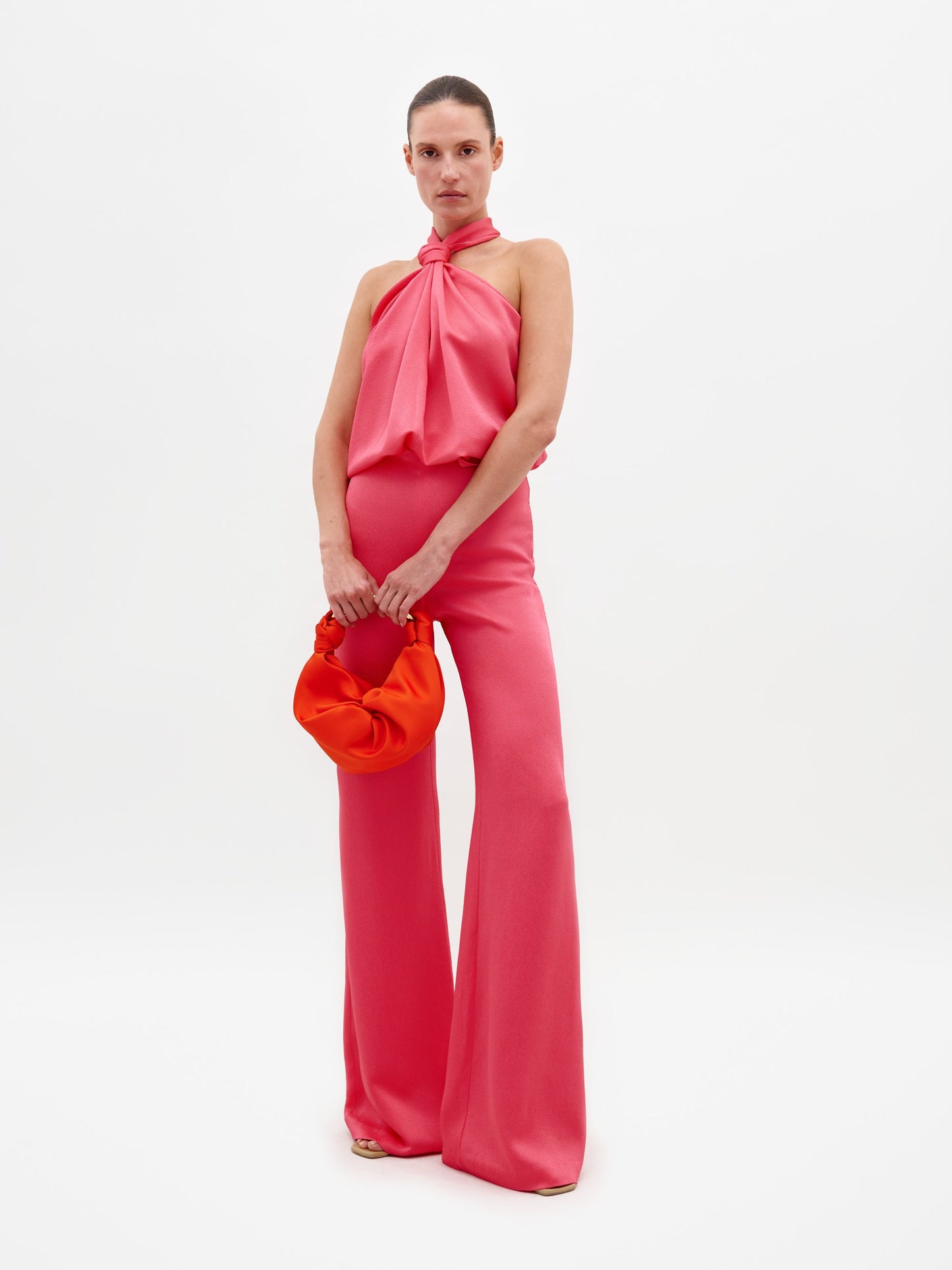 A person wearing a Palermo Pant Peony set, which includes a pink halter top and matching wide-leg pants, holds an orange handbag against a plain white background. This stylish ensemble is available for pre-order, with a ship by date of February 15th, 2025.