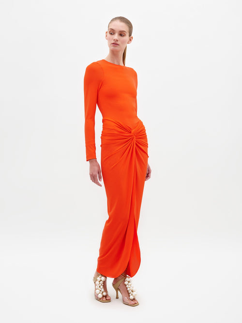 A person wearing the Sydelle Dress Tangerine, a long-sleeved orange dress featuring a knotted detail in the front, stands looking to the side. They are also donning sandals embellished with pearl-like beads. This elegant outfit will be available for pre-order and is anticipated to ship by 2025.