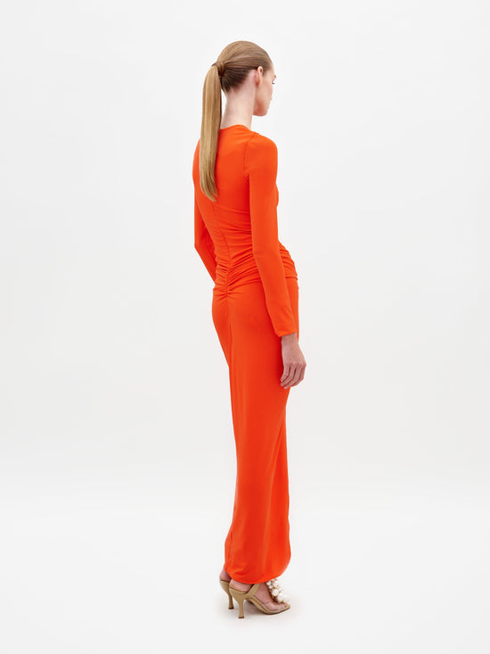 A person wearing the Sydelle Dress Tangerine, a long-sleeved orange dress featuring a knotted detail in the front, stands looking to the side. They are also donning sandals embellished with pearl-like beads. This elegant outfit will be available for pre-order and is anticipated to ship by 2025.