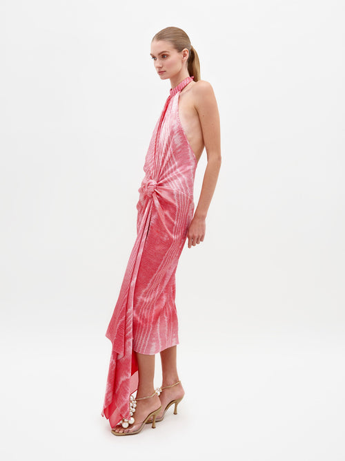 A person wearing Theodora Dress Peony Moire, a sleeveless, tie-dyed pink dress with a high neckline and front knot detail, paired with beige high-heeled sandals. Pre-order now to ensure you receive it by the ship-by date of February 15th, 2025.