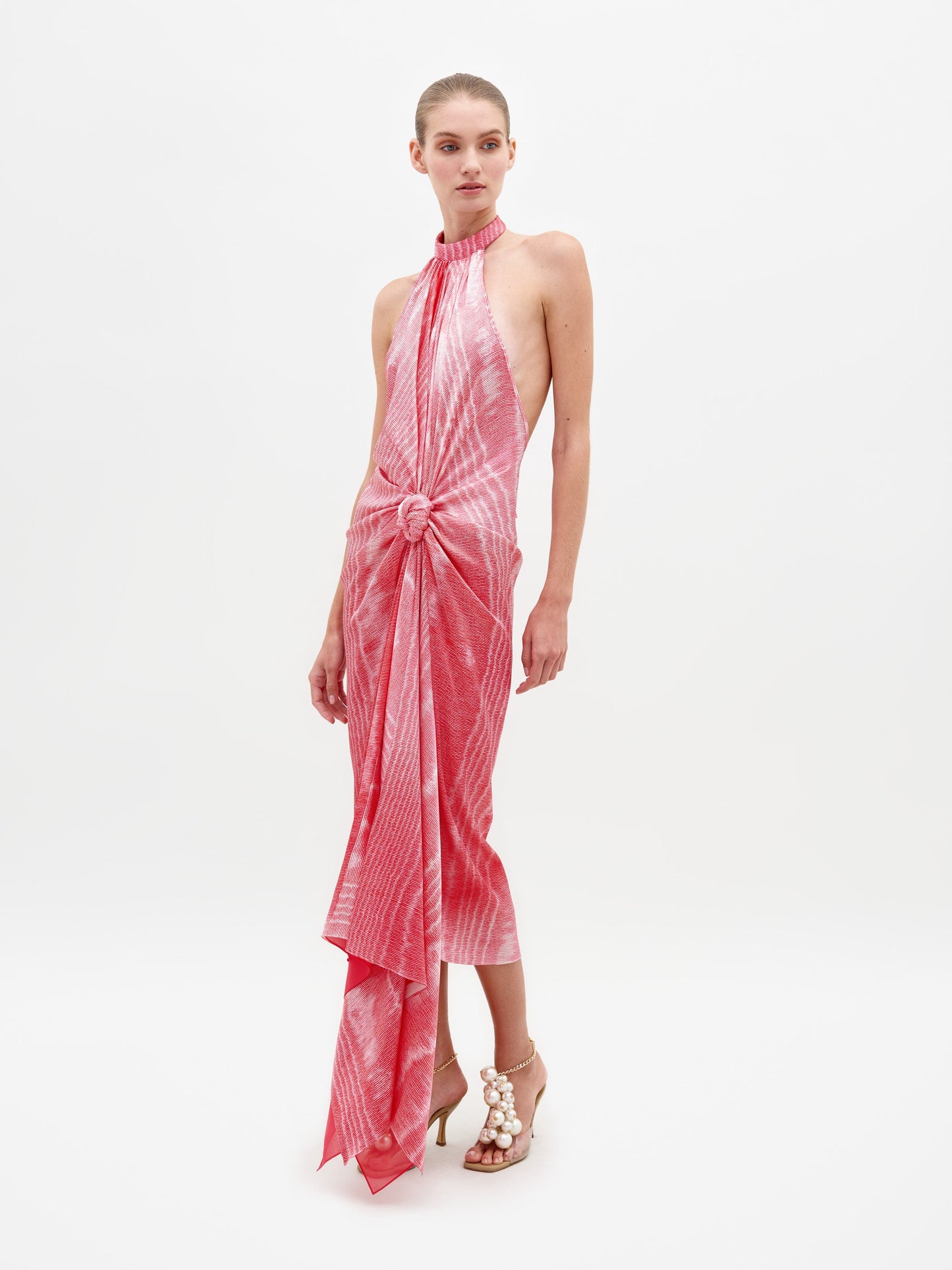 A person wearing Theodora Dress Peony Moire, a sleeveless, tie-dyed pink dress with a high neckline and front knot detail, paired with beige high-heeled sandals. Pre-order now to ensure you receive it by the ship-by date of February 15th, 2025.