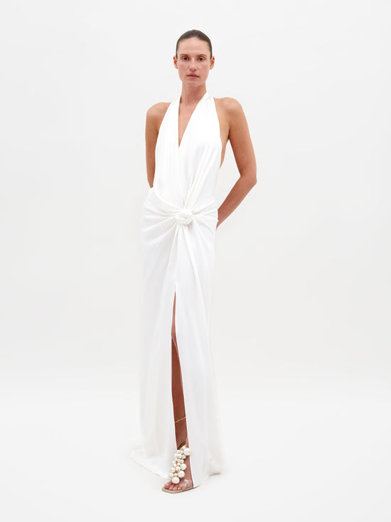 Person standing against a white background, wearing the Torgiano Dress White, which features a sleeveless halter design with a knot detail at the waist and a front slit. Pre-order now to ensure your dress is shipped by February 15th, 2025.