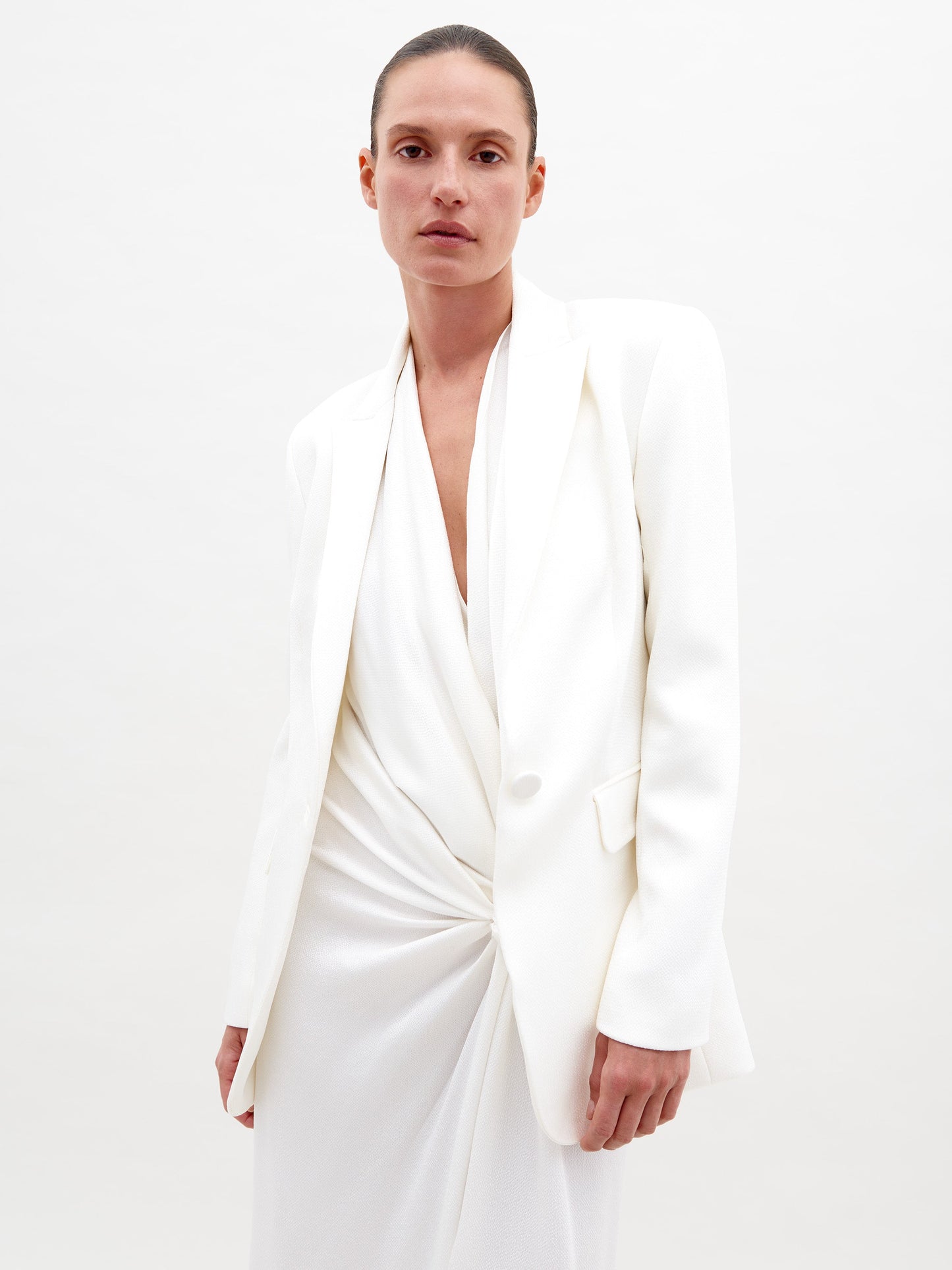 A person with a neutral expression is wearing the Rebeca Jacket White and white pants, standing against a plain background with arms crossed, available for pre-order starting February 15th, 2025.