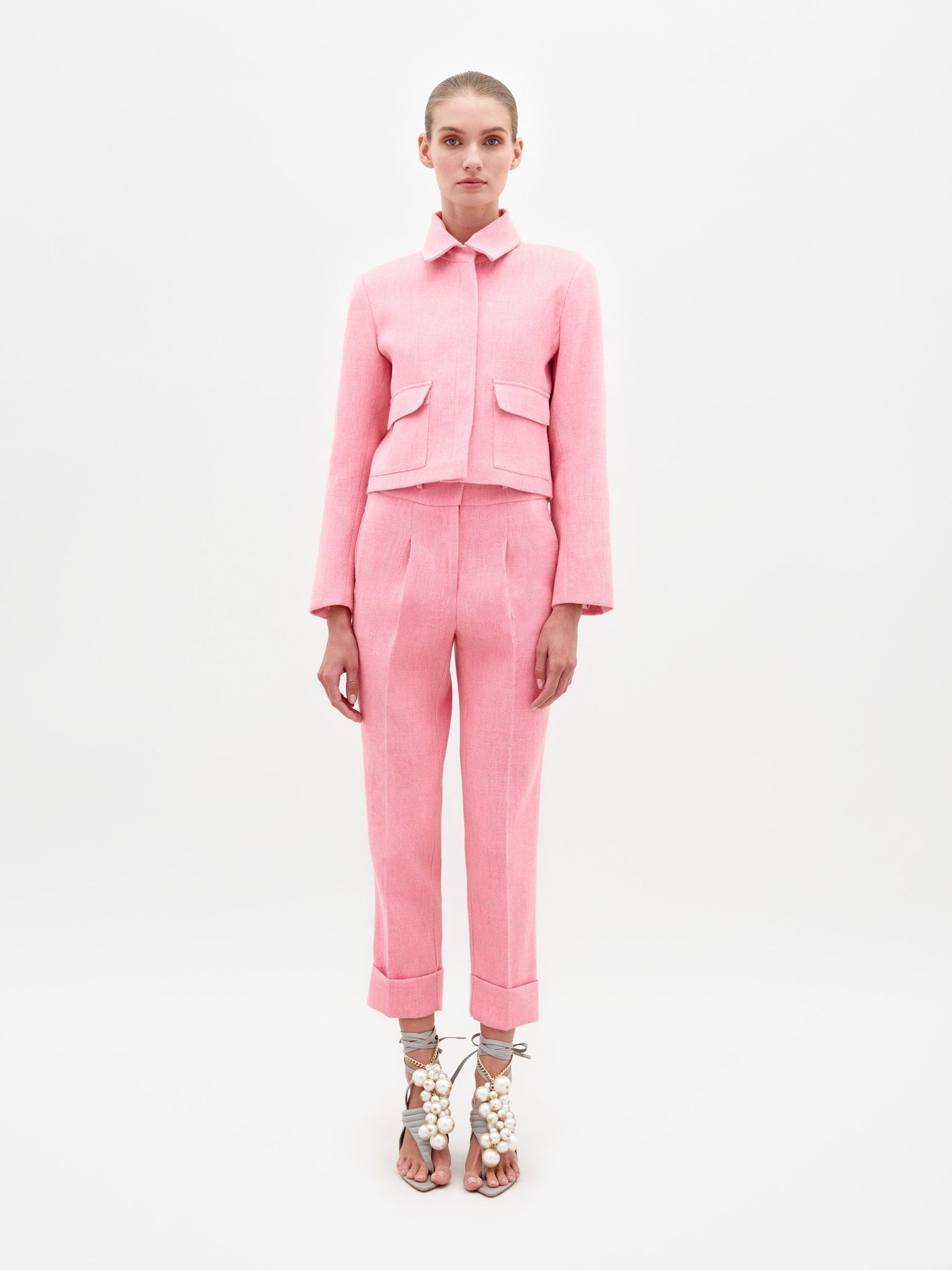 A person stands against a plain background, wearing the Moad Pant Pink suit, featuring a tailored cropped jacket and matching pink pants. They complete their look with high-heeled shoes adorned with white embellishments, ready for shipment by February 15th, 2025.