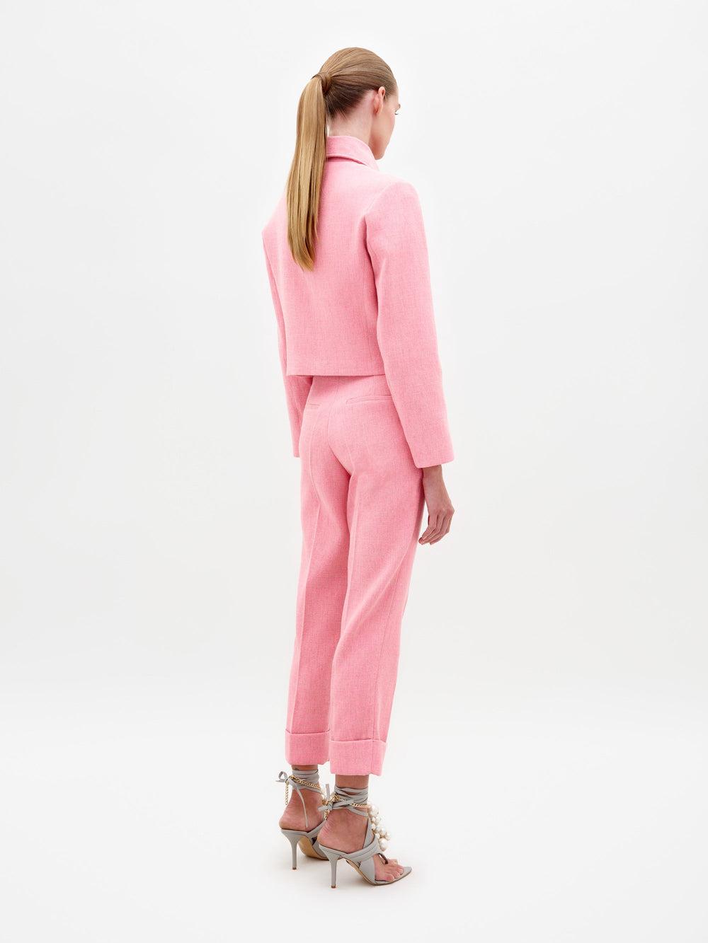 A person stands against a plain background, wearing the Moad Pant Pink suit, featuring a tailored cropped jacket and matching pink pants. They complete their look with high-heeled shoes adorned with white embellishments, ready for shipment by February 15th, 2025.
