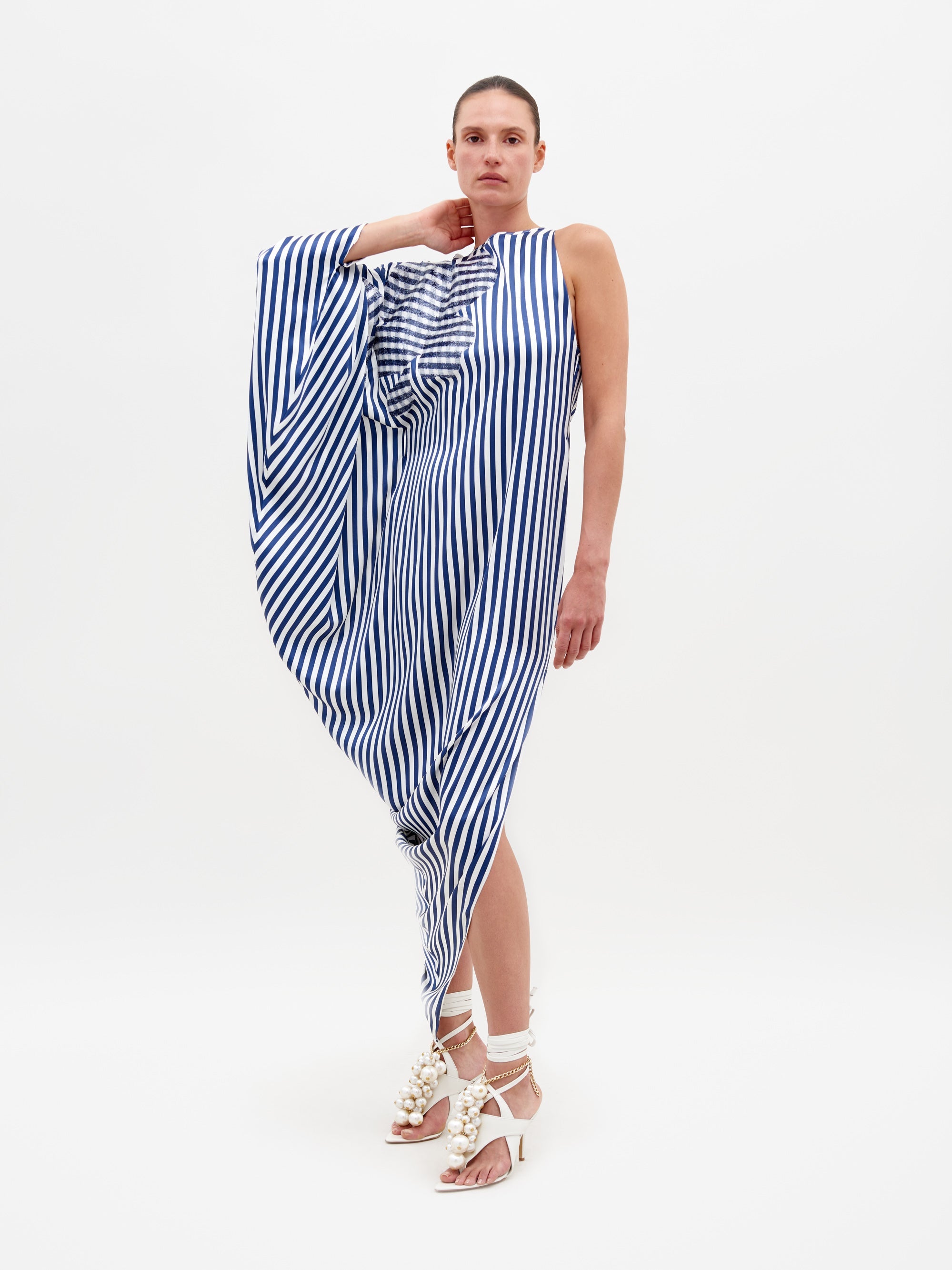 Person wearing the Golda Dress in White and Blue Stripes, an asymmetrical design with draped fabric, paired with white strappy sandals featuring embellishments, standing against a plain white background. Pre-order now to ensure it ships by February 15th, 2025.