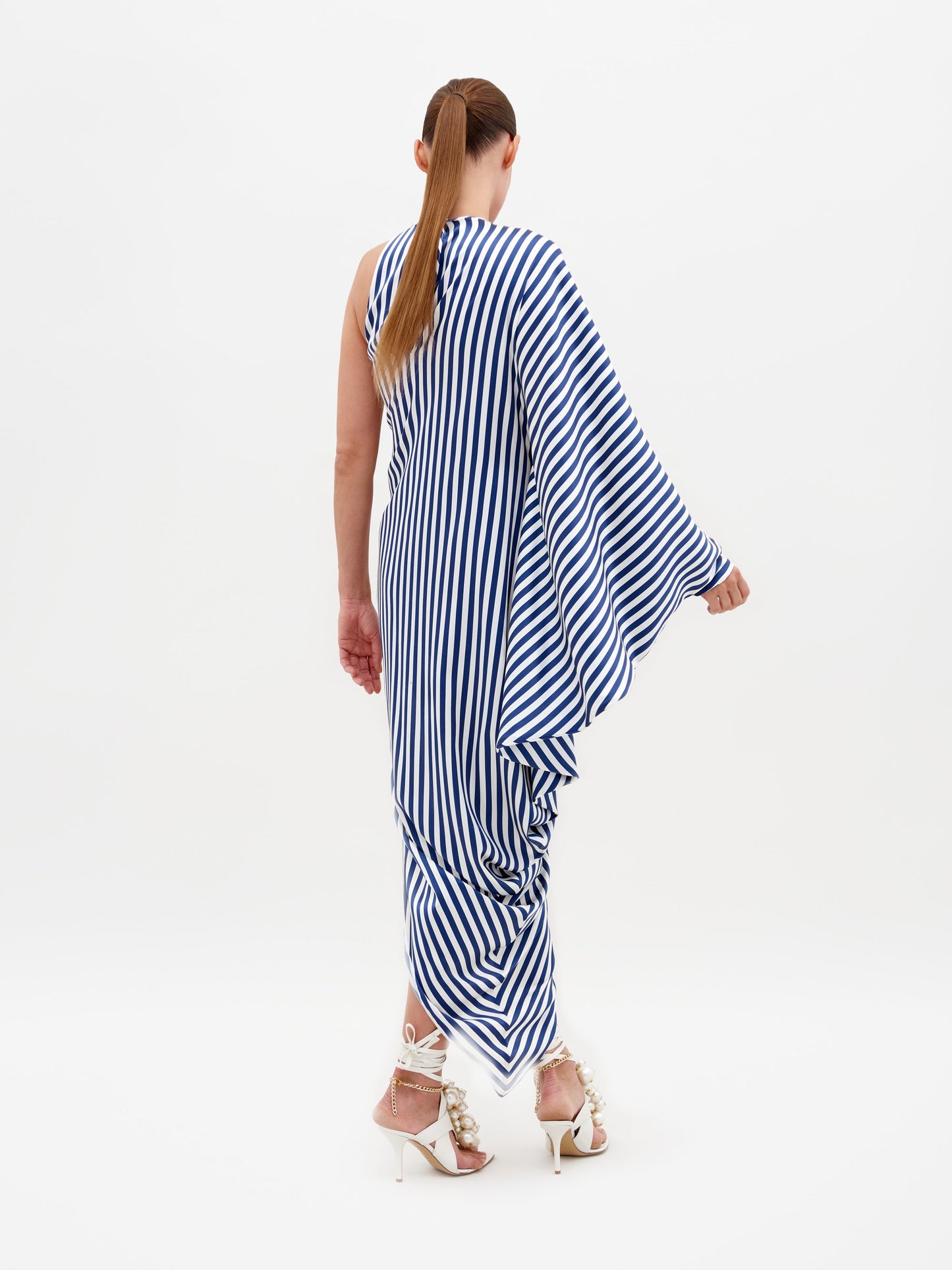 Person wearing the Golda Dress in White and Blue Stripes, an asymmetrical design with draped fabric, paired with white strappy sandals featuring embellishments, standing against a plain white background. Pre-order now to ensure it ships by February 15th, 2025.