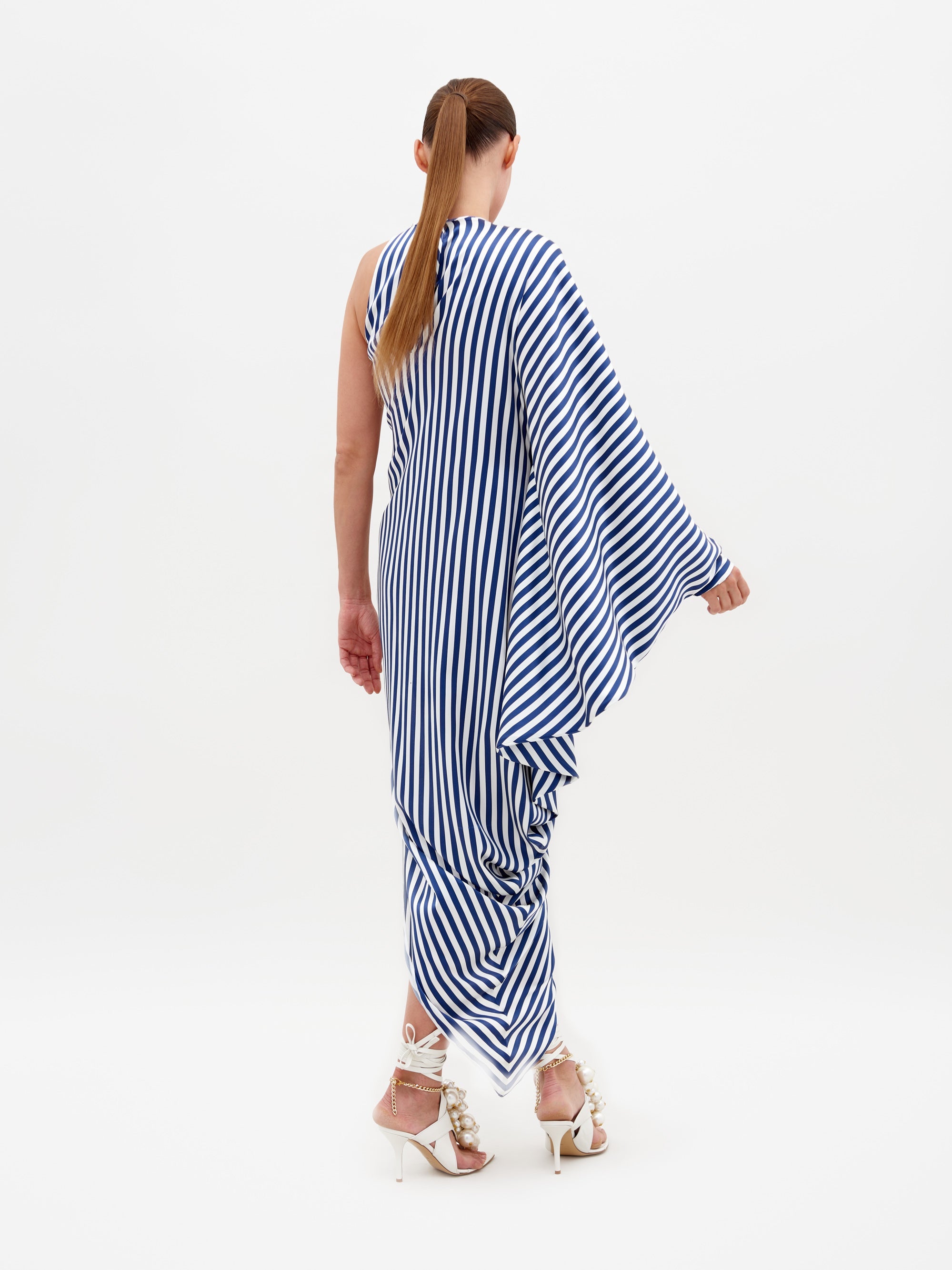 Person wearing the Golda Dress in White and Blue Stripes, an asymmetrical design with draped fabric, paired with white strappy sandals featuring embellishments, standing against a plain white background. Pre-order now to ensure it ships by February 15th, 2025.