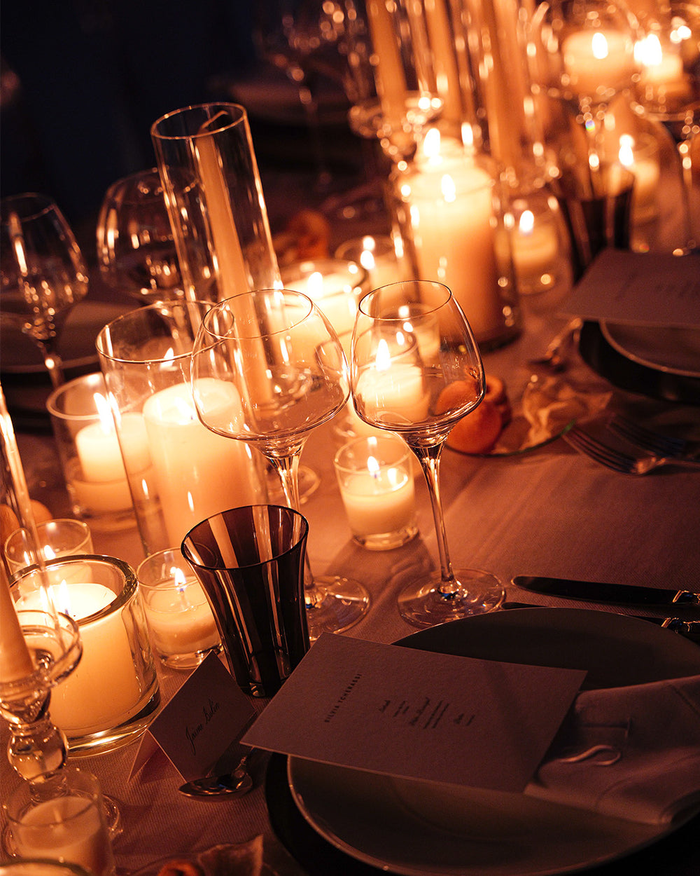 A warmly lit dining table setting with lit candles in glass holders, wine glasses, plates, napkins, and silverware in a cozy ambient scene.