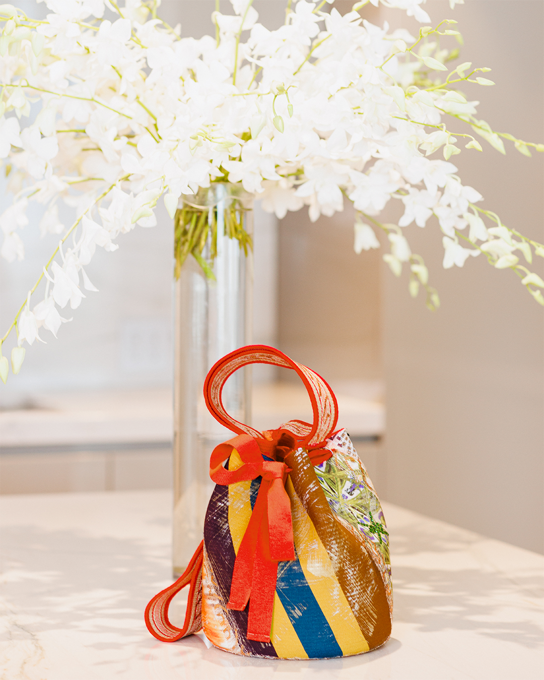 A colorful, cylindrical bag with red handles sits on a marble surface in front of a tall vase with white flowers.