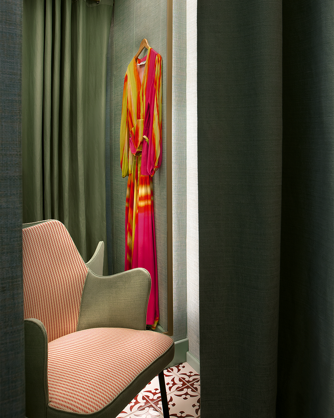 A pink and yellow dress hangs on a wall in a fitting room with dark green curtains and a beige chair.