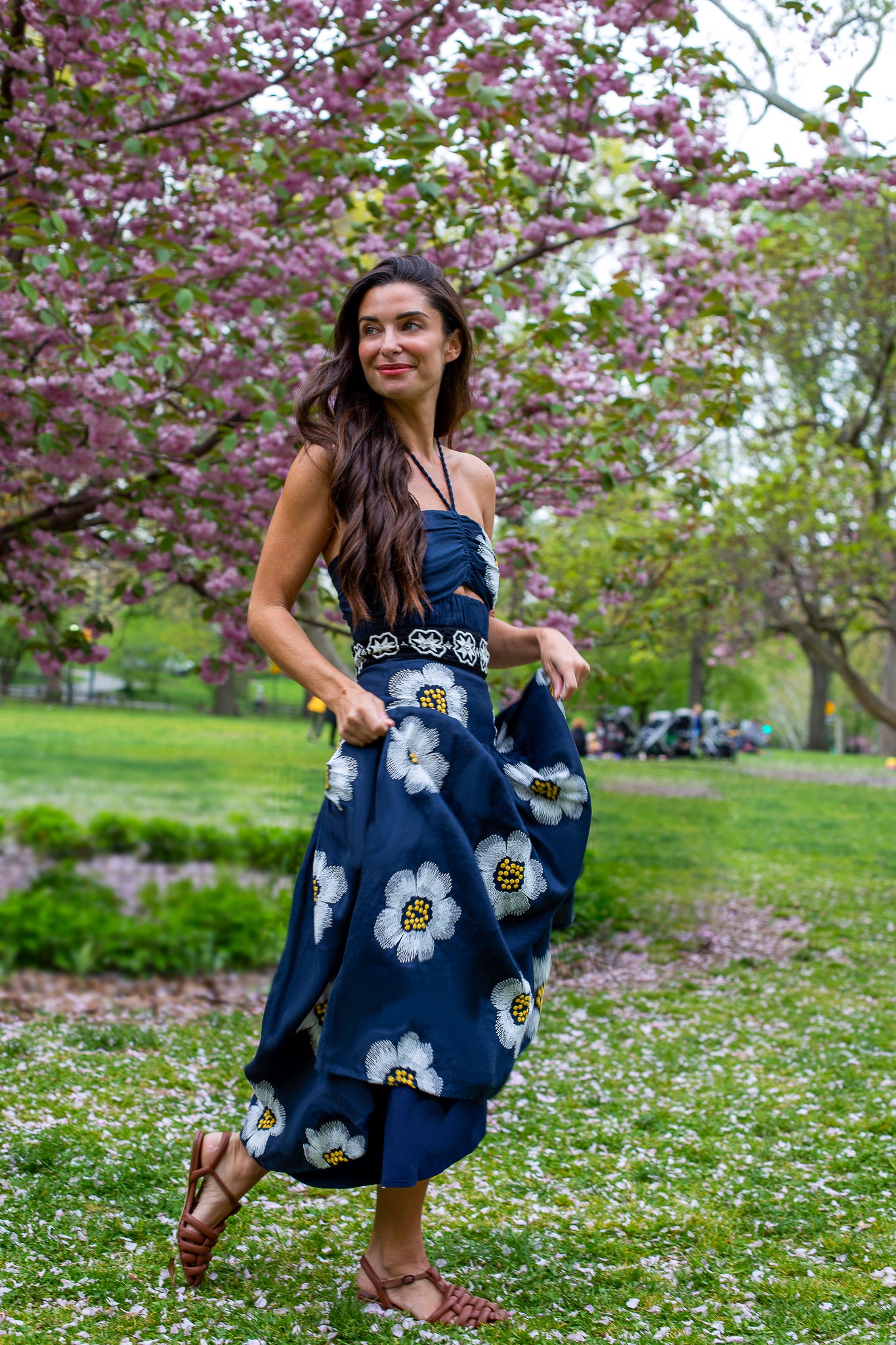 A woman in a floral dress stands in a park with blooming trees, walking on grass while holding her skirt.