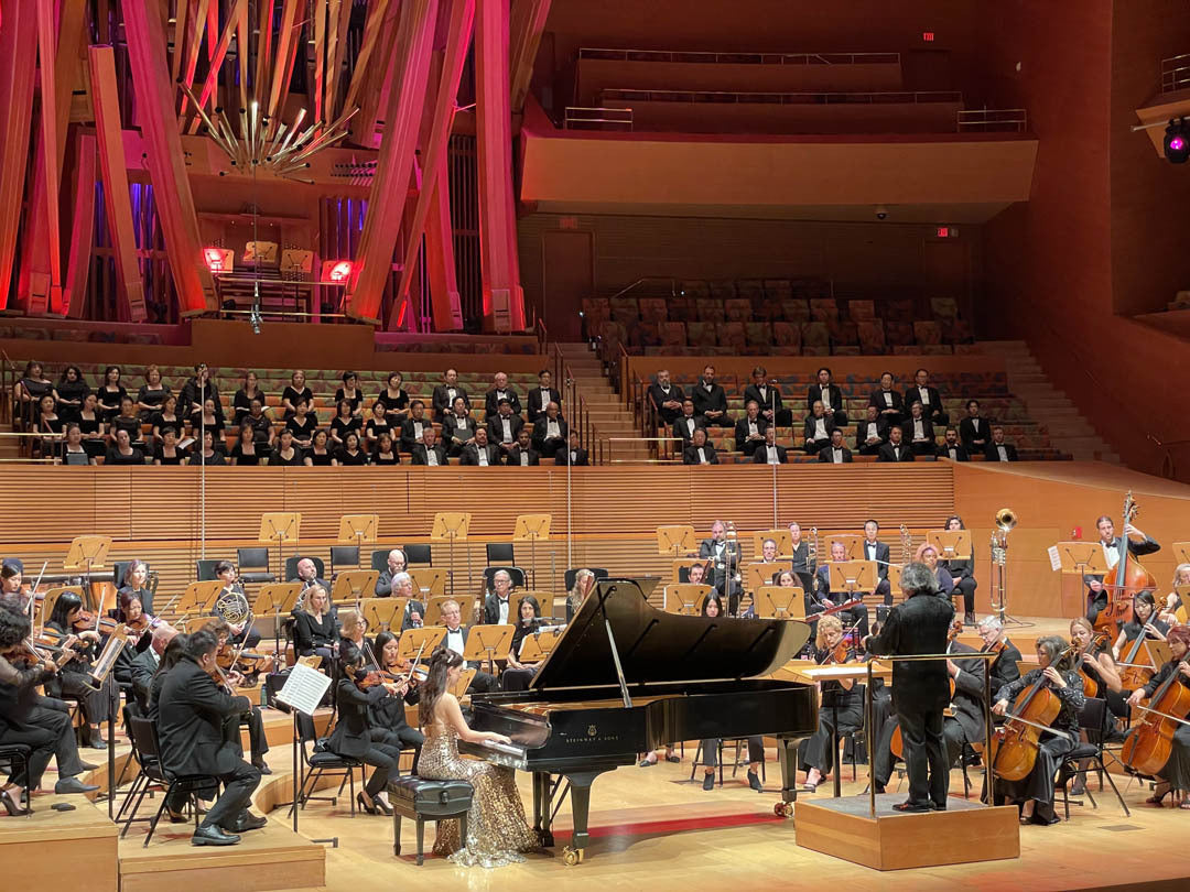 A pianist performs with an orchestra on stage in a concert hall. The choir is seated in the background. The hall is lit with red and orange lights.