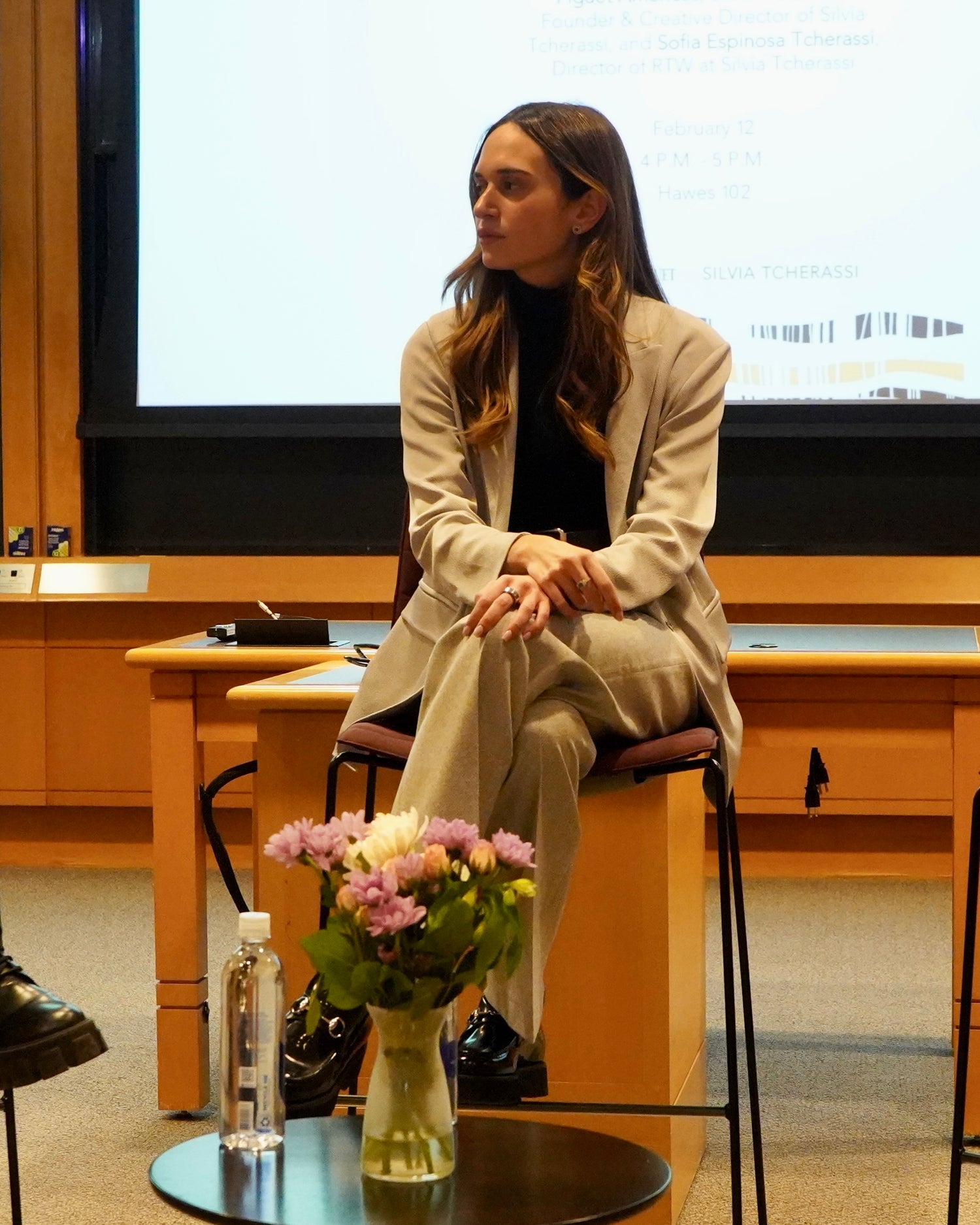 A woman in a beige suit sits on a desk chair in a lecture room with a presentation slide in the background. A table with flowers and a water bottle is in the foreground.