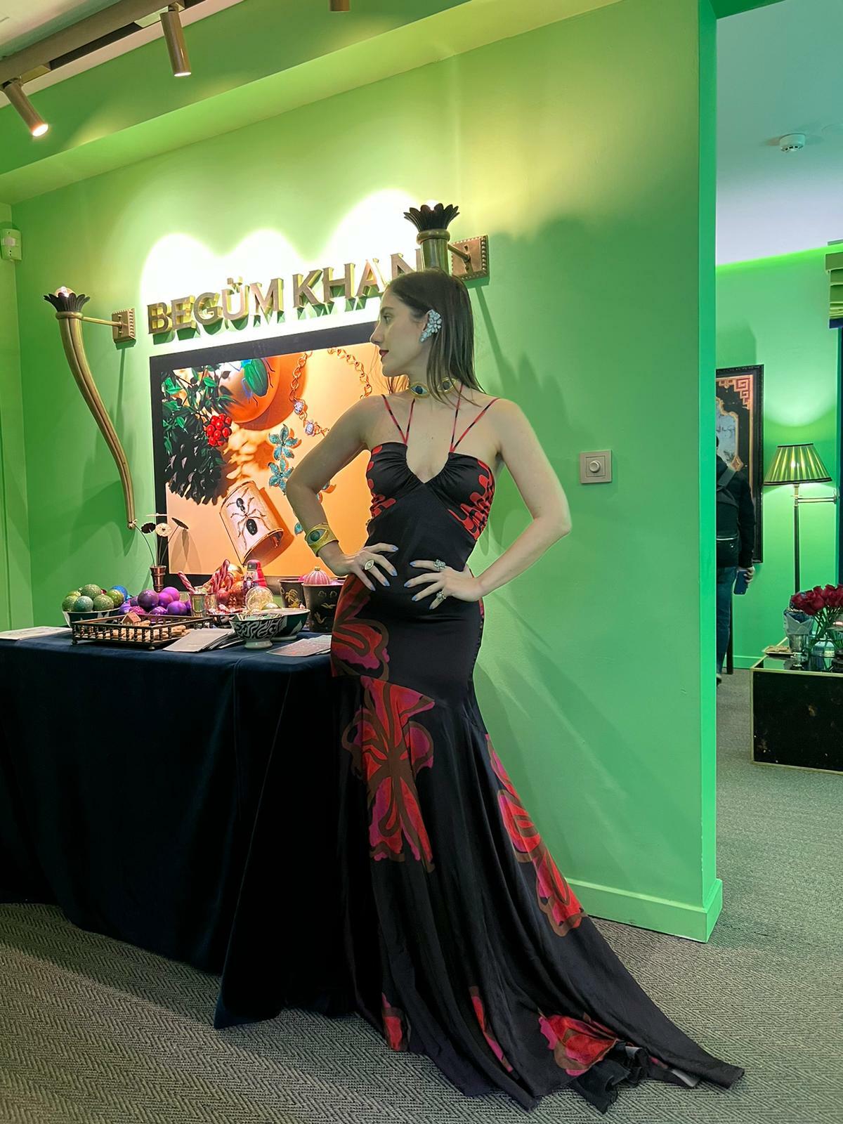 A woman in a black and red dress stands next to a table in a room with green walls and signage reading "BEGUM KHAN.