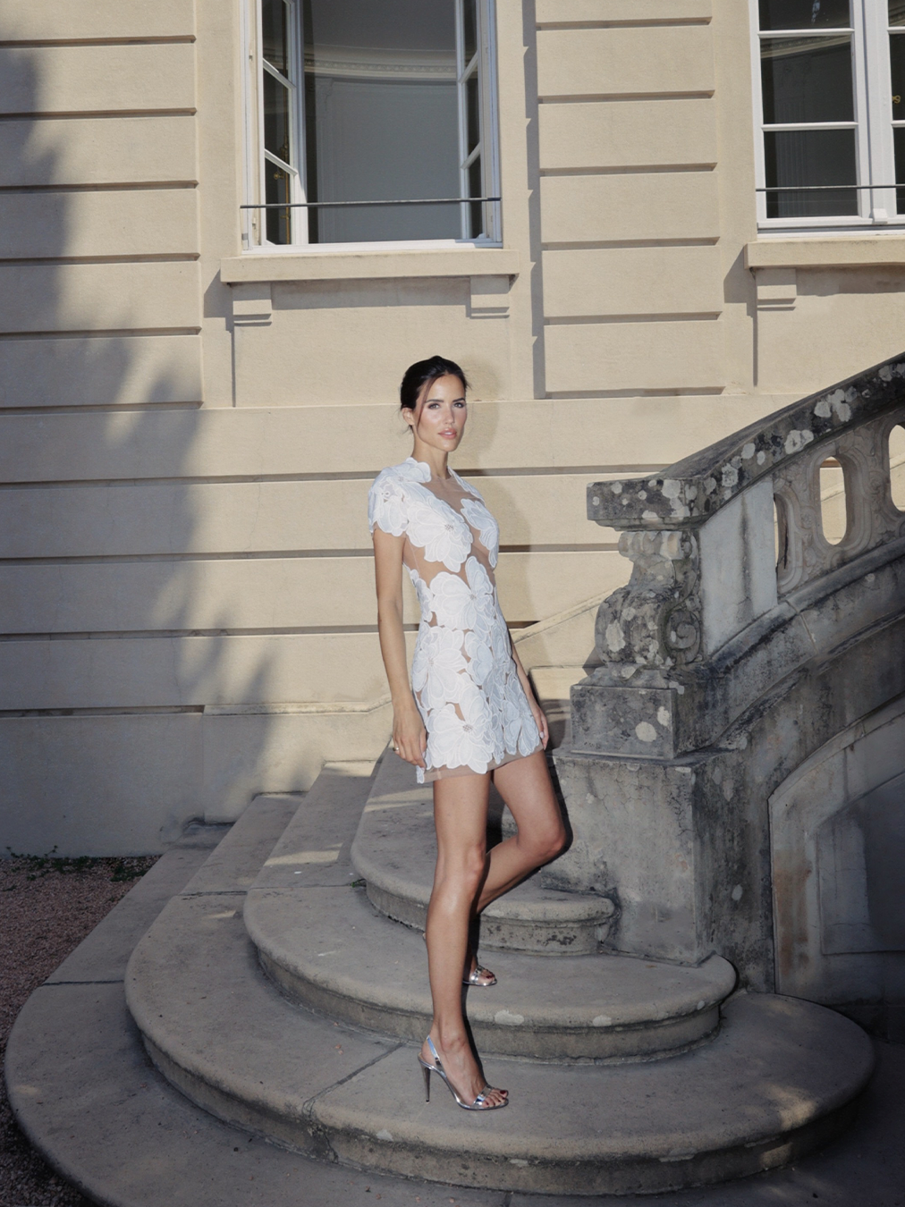 A woman in a short white lace dress stands on curved stone steps in front of a beige building with large windows.