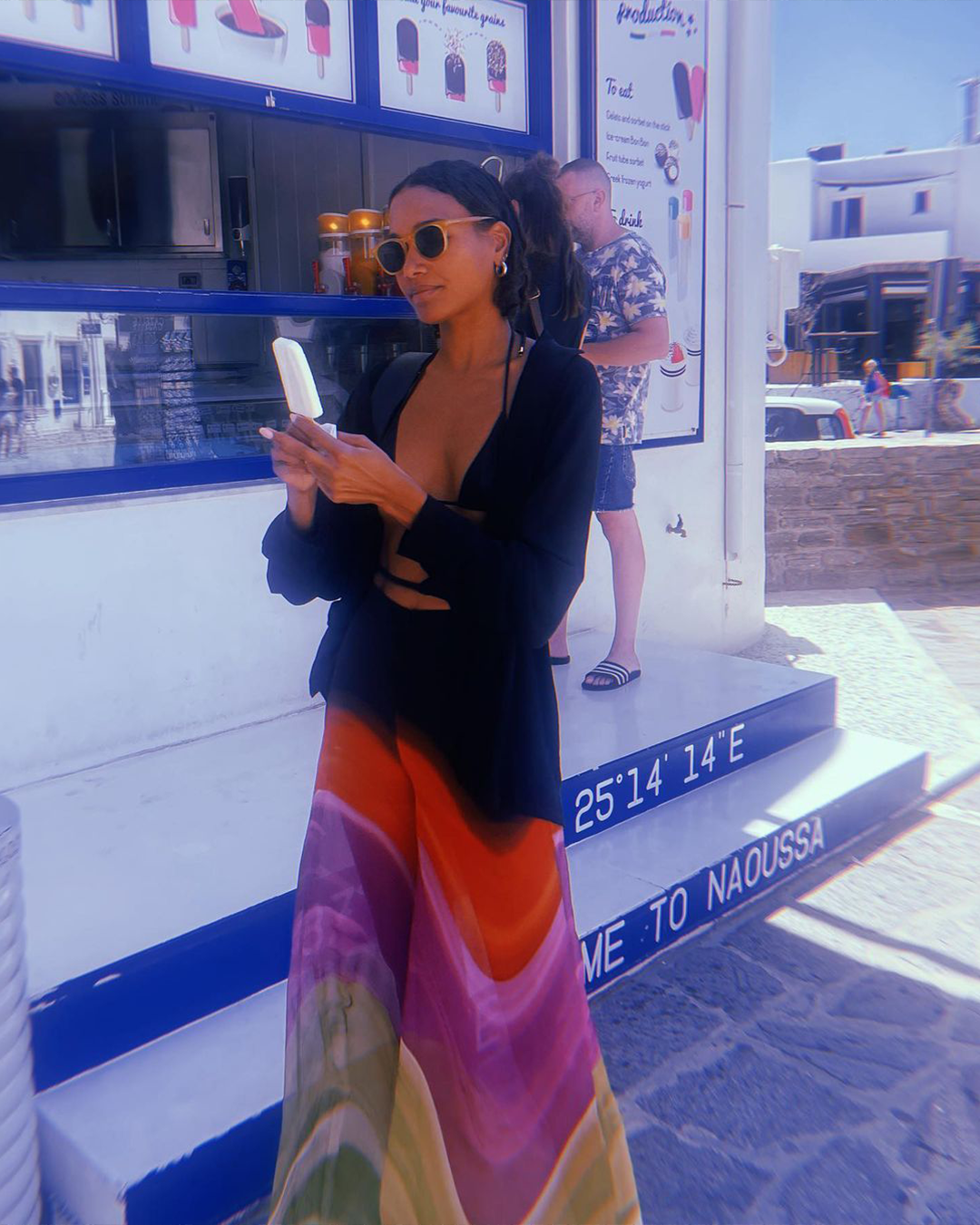 A woman in sunglasses holds an ice cream cone. She wears a colorful skirt and stands outside a shop with a blue and white facade.