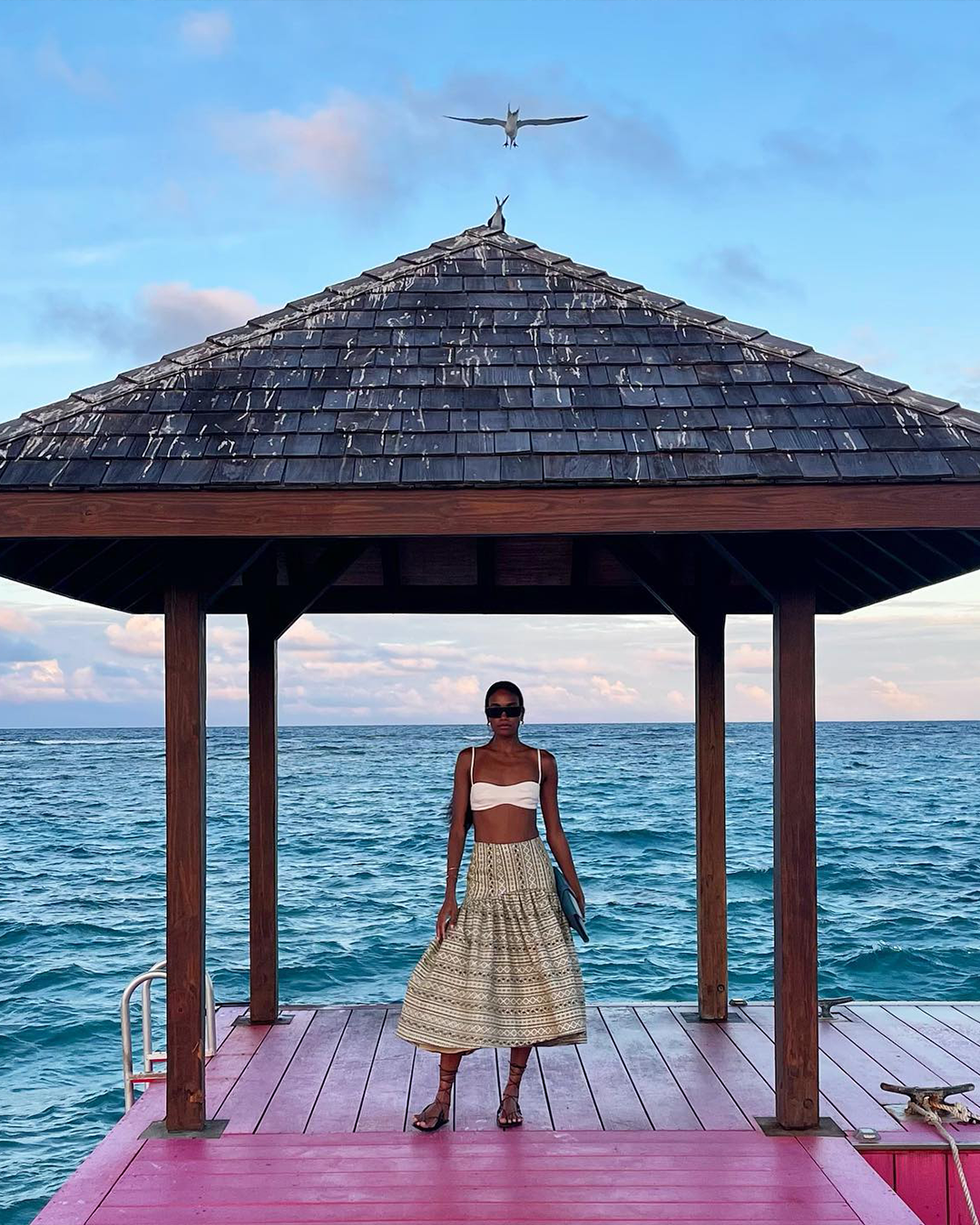 A person stands on a dock under a wooden gazebo by the ocean, with a bird flying overhead.