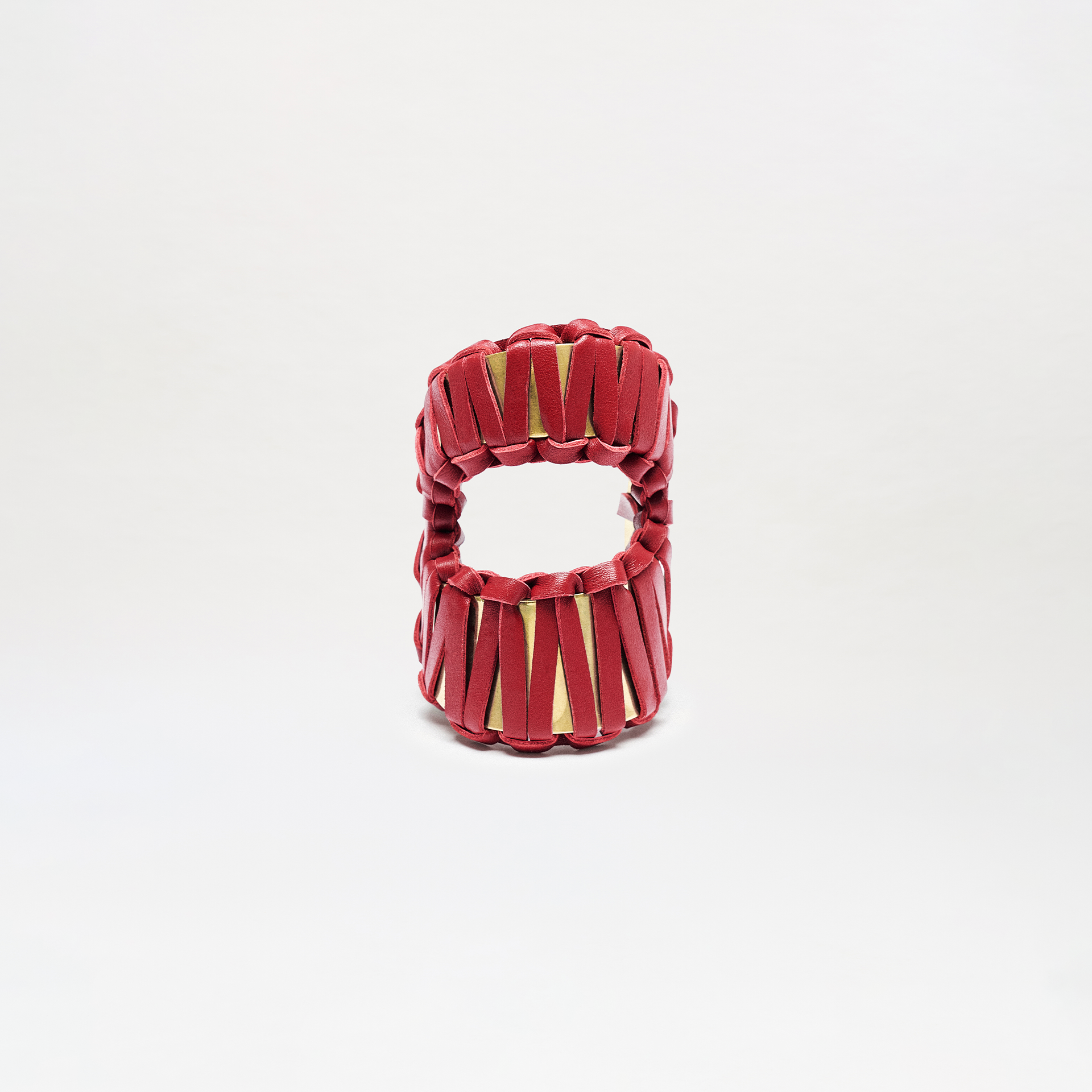 The Majel Cuff is a cylindrical accessory with a hollow center, featuring vertical red bands that crisscross and reveal a gold interior. It is displayed against a plain white background.
