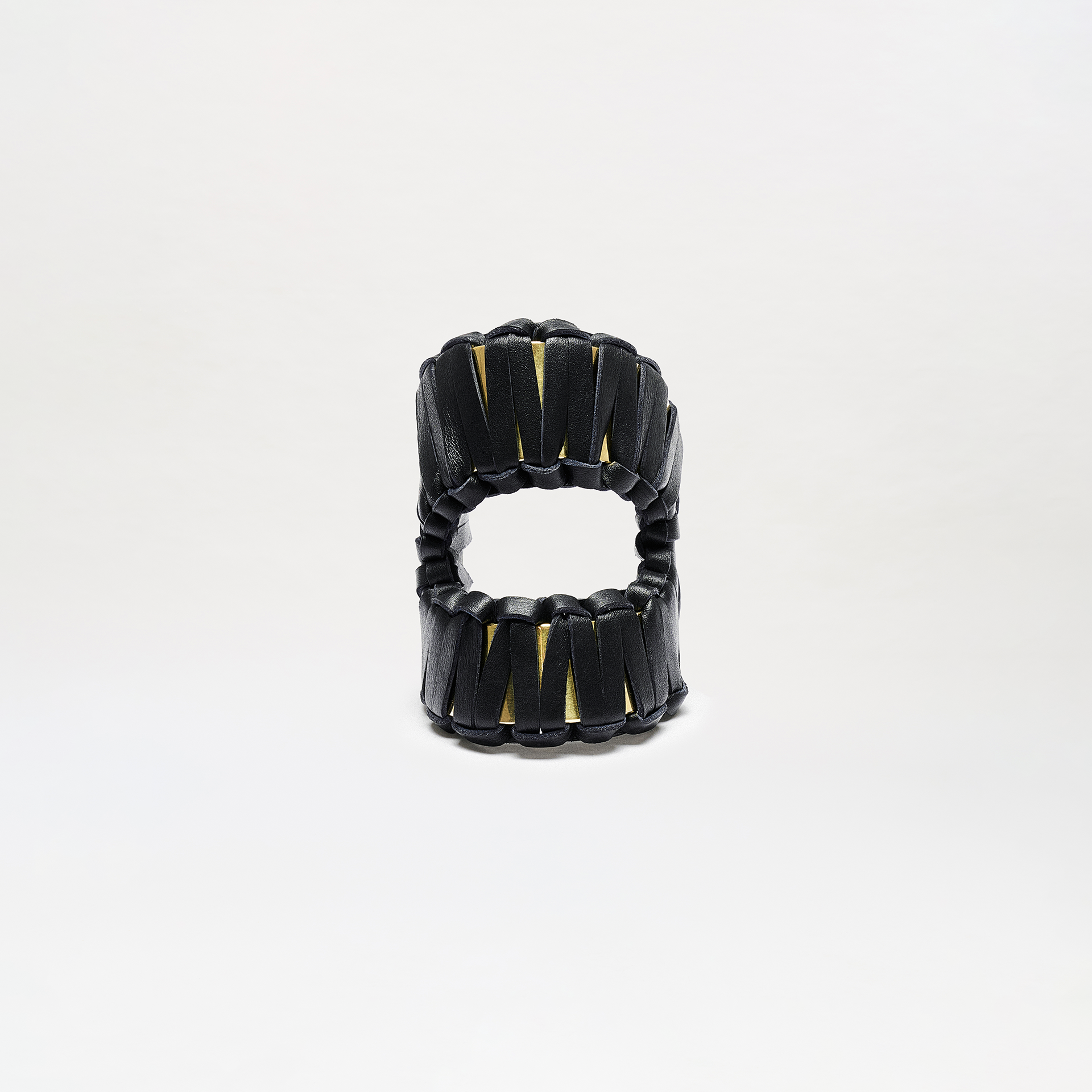 The Majel Cuff is a sculptural black accessory with vertical slits revealing a gold interior, shaped like two stacked rings with an open center, shown against a plain white background.