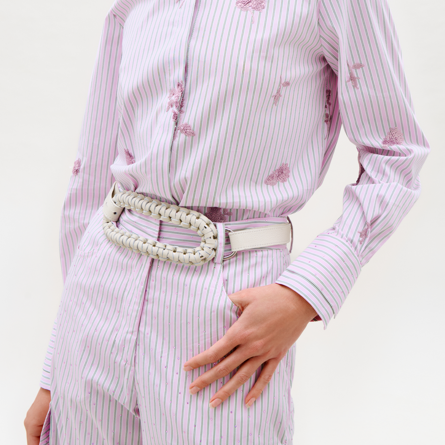 A person stands against a plain background, wearing a light purple pinstripe shirt and matching floral-detail pants, accessorized with the Cloris Belt in white and resting one hand on their hip.