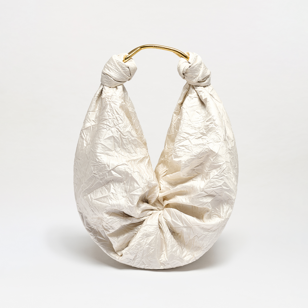 The Sofia Bag (Maxi) features a white crinkled texture, rounded shape, gathered Italian fabric, and two top knots joined by a shiny gold-toned handle. It is displayed against a plain light background.