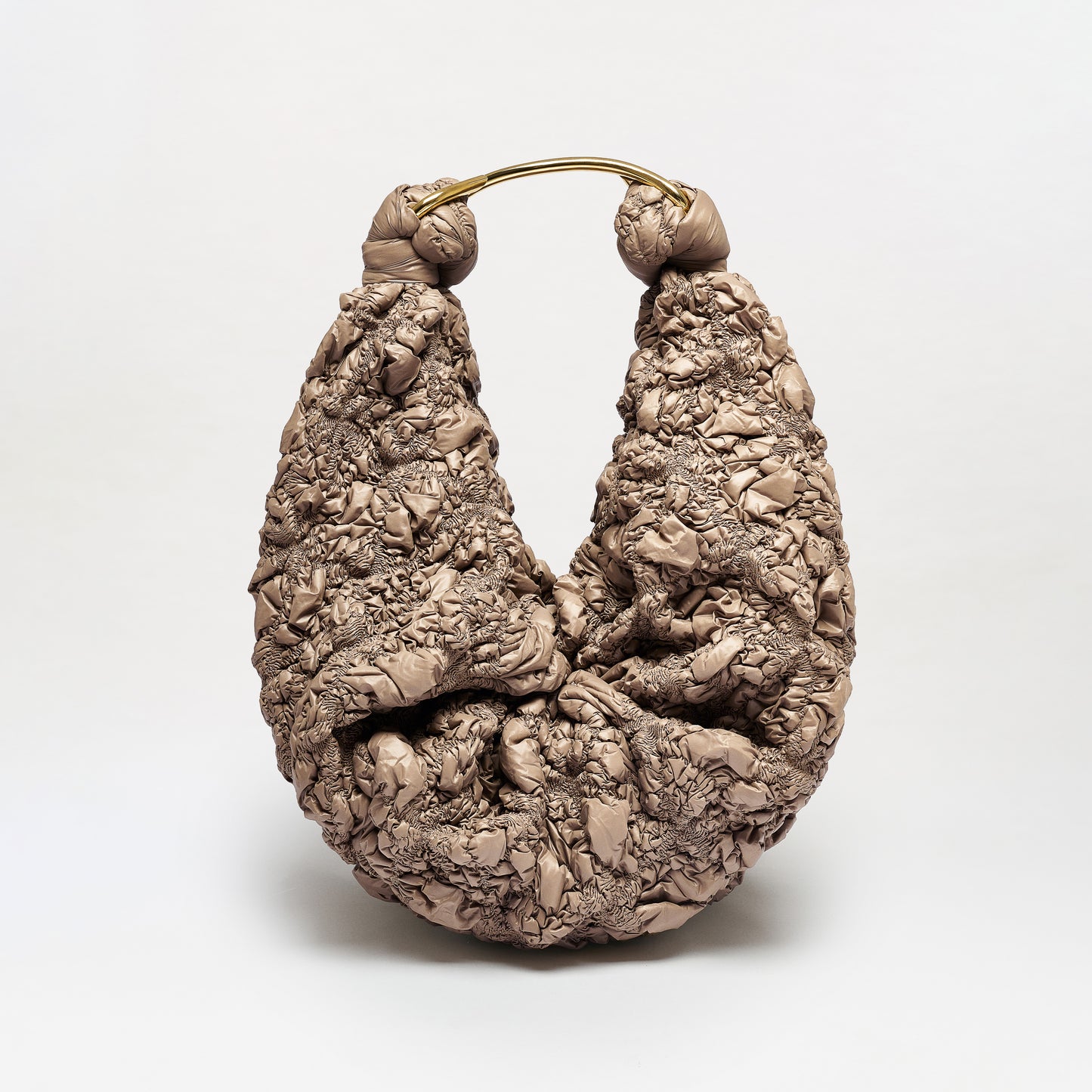 The Sofia Bag (Maxi) is a sculptural, crescent-shaped bag crafted from crinkled taupe Italian fabric with a curved gold-toned handle, shown standing upright against a plain white background.