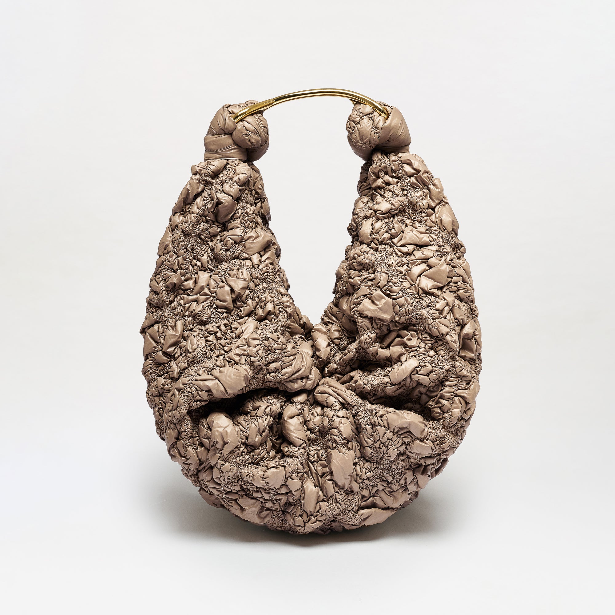 The Sofia Bag (Maxi) is a sculptural, crescent-shaped bag crafted from crinkled taupe Italian fabric with a curved gold-toned handle, shown standing upright against a plain white background.
