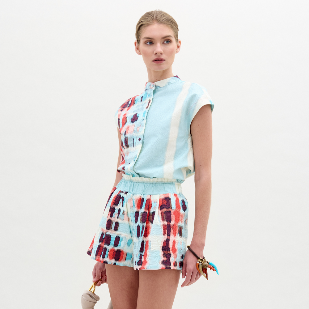 A woman wears the Adele Blouse, a collarless light blue top with short sleeves, paired with matching high-waisted shorts in Italian fabric featuring a multicolored abstract pattern, standing against a plain white background.