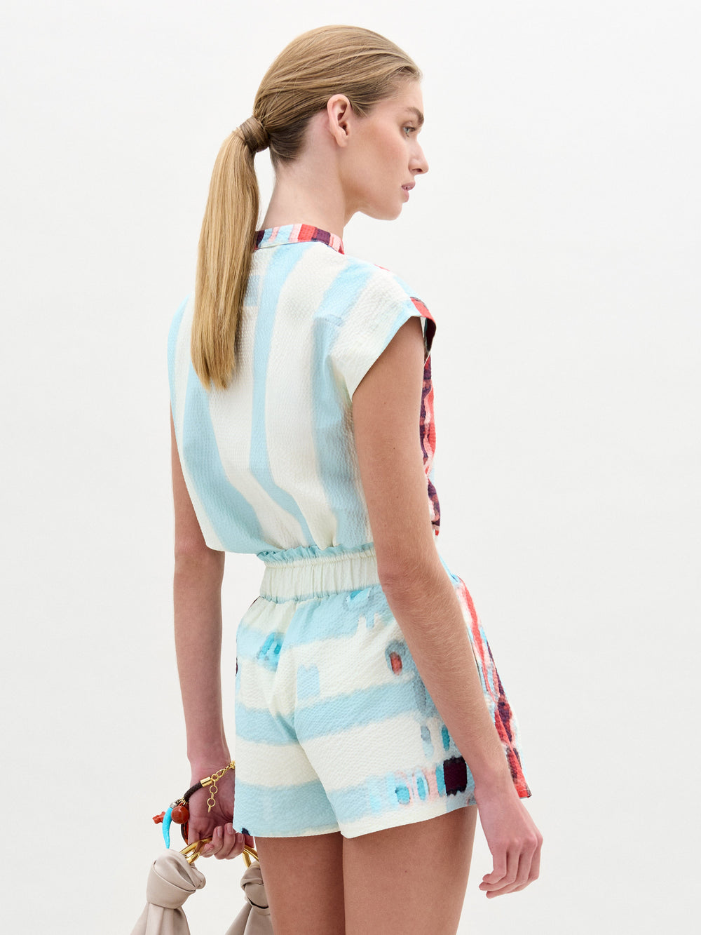 A woman wears the Adele Blouse, a collarless light blue top with short sleeves, paired with matching high-waisted shorts in Italian fabric featuring a multicolored abstract pattern, standing against a plain white background.