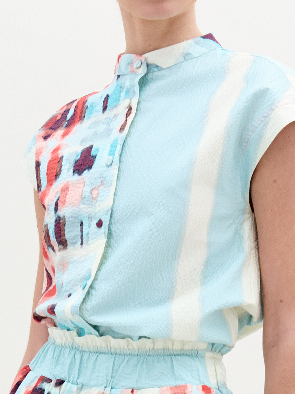 A person wearing the Adele Blouse—a light blue, short-sleeved top made in Colombia with a red, purple, and white abstract pattern, high collar, and fabric-covered buttons.